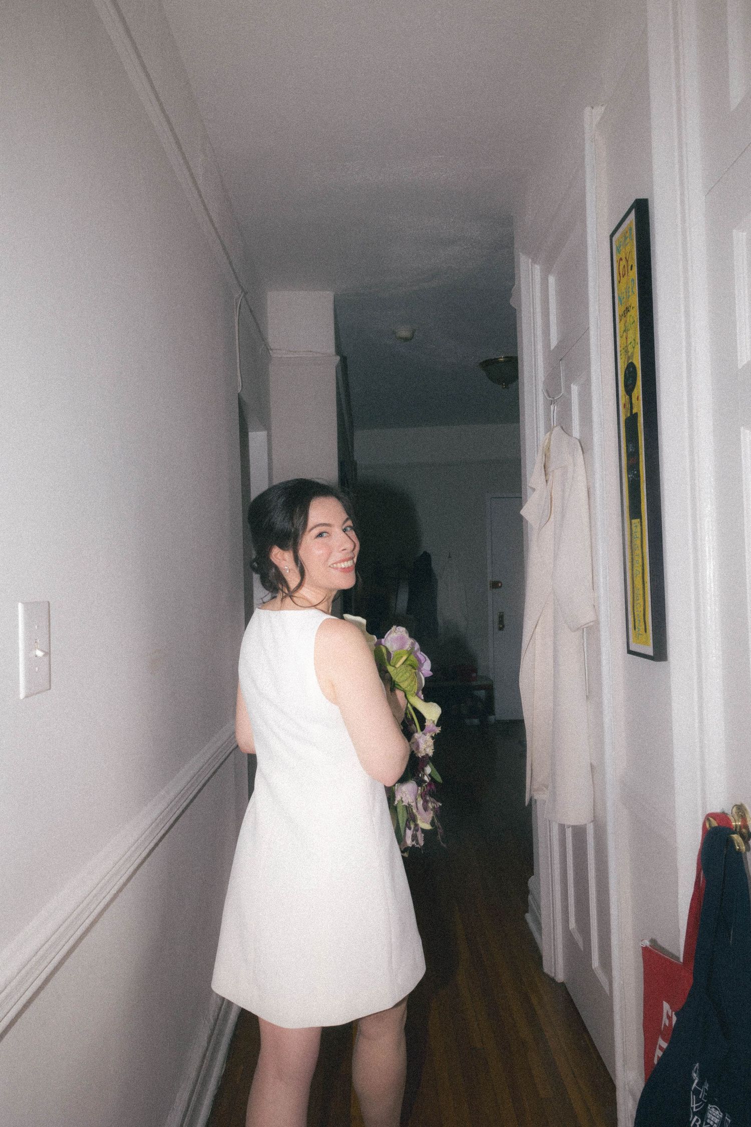 Bride in white mini dress holds colorful bouquet in apartment hallway, smiling over shoulder.