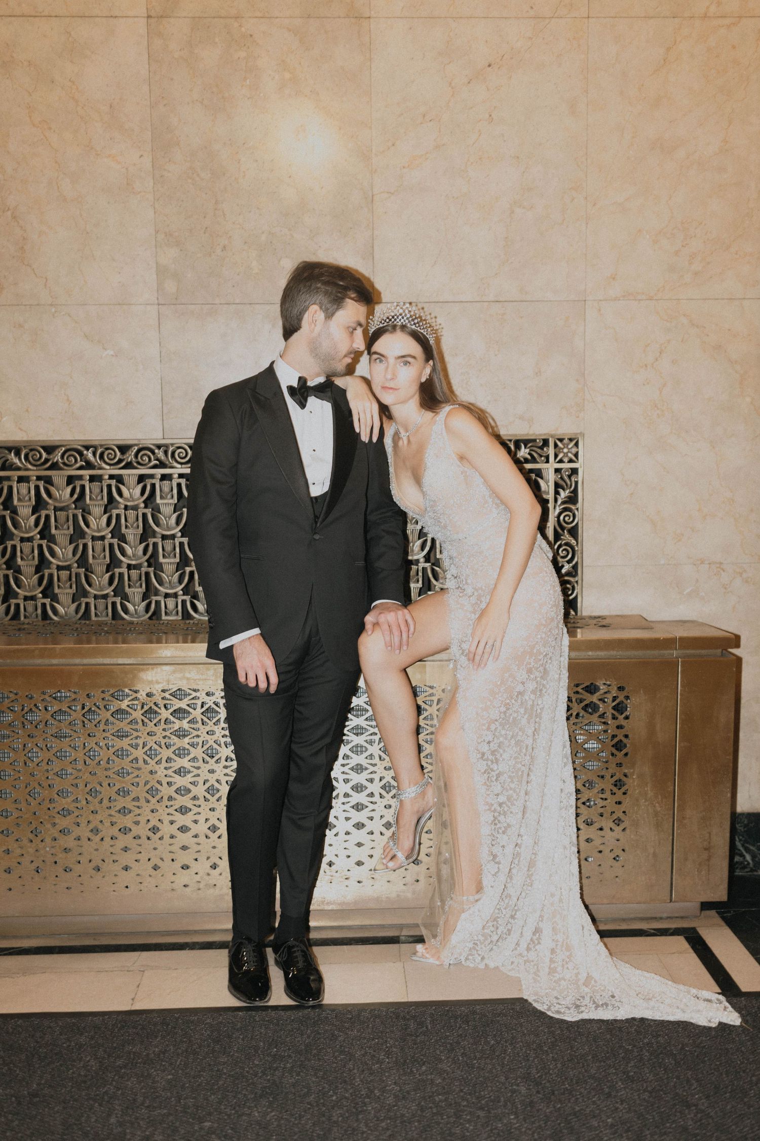 Groom in black tuxedo and bride in lace gown with crystal tiara pose elegantly in a marble-walled venue.