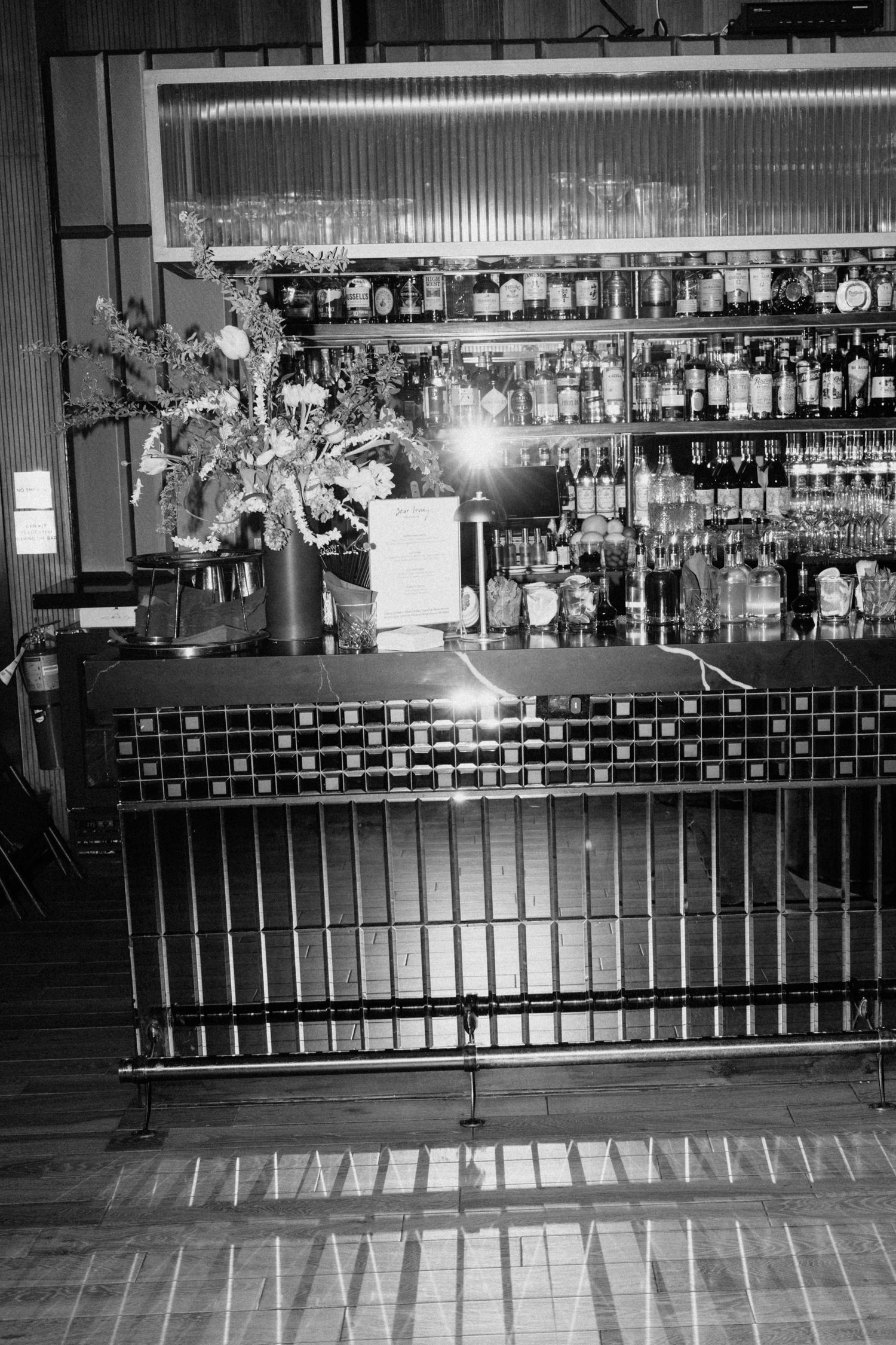 Black and white photo of an elegant bar with floral arrangement and shelves of bottles.