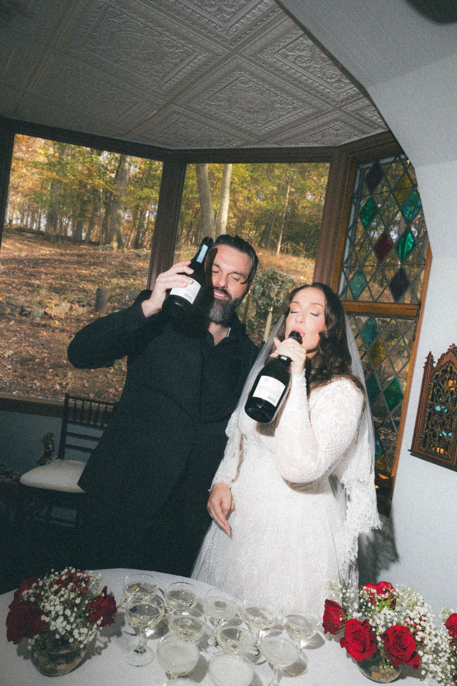 Bride and groom drinking champagne from bottles near stained glass bay window at wedding reception.