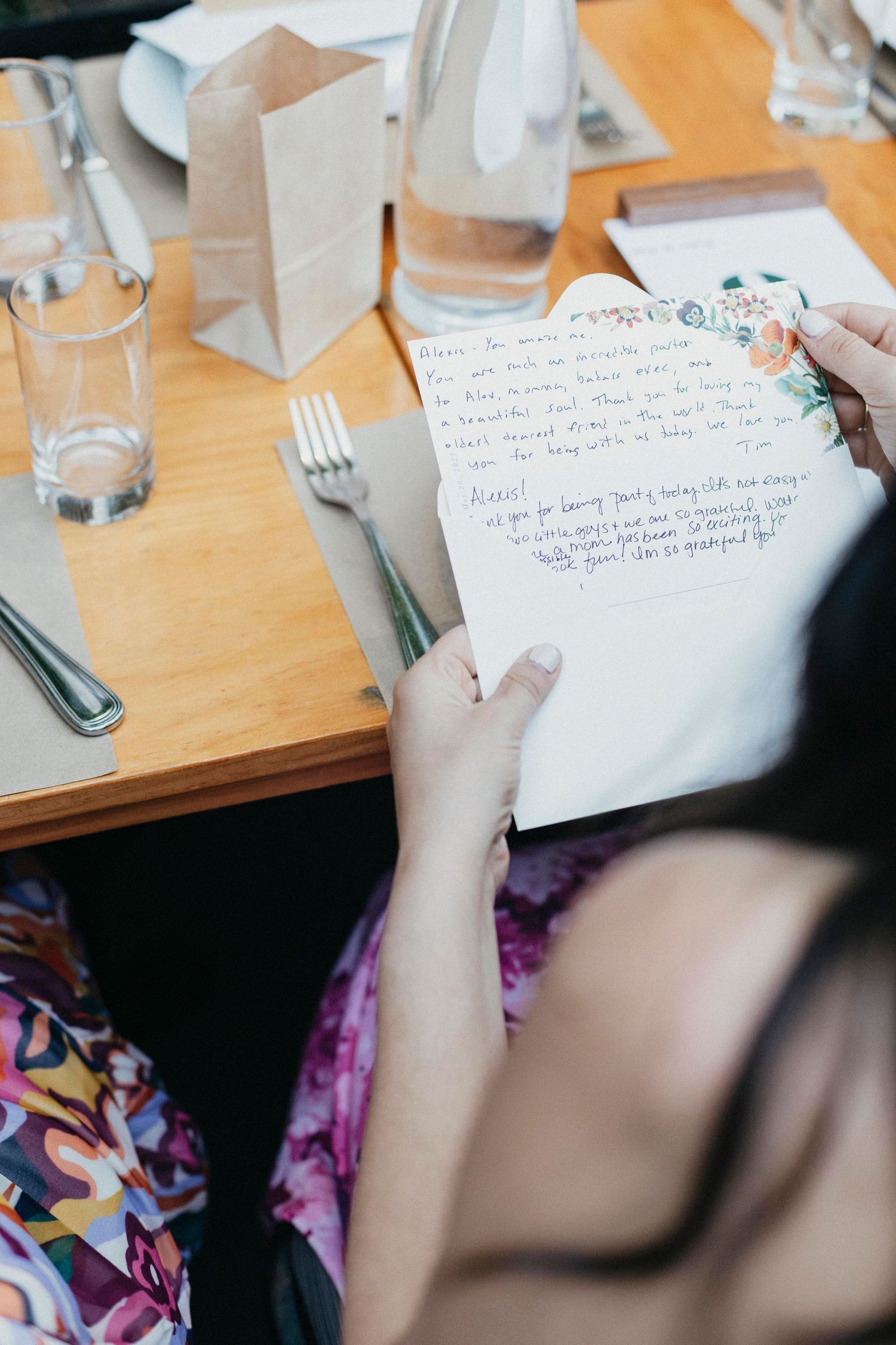 Guest reads handwritten floral wedding card at wooden reception table with wine glasses and place settings.