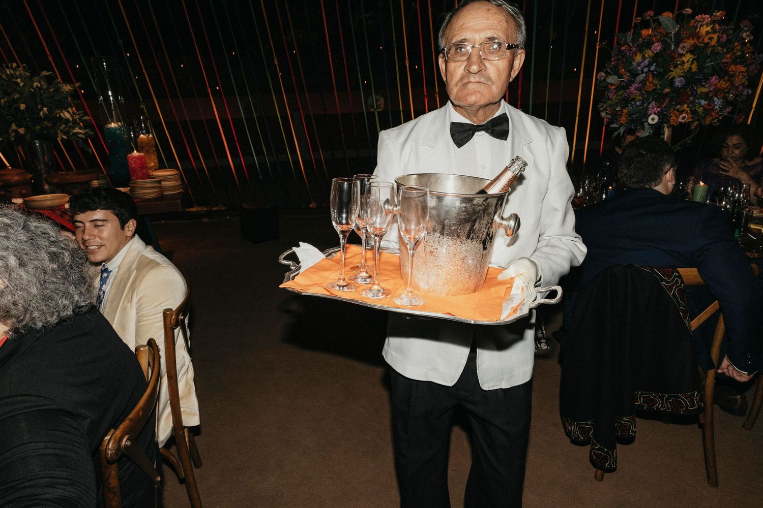 Formal waiter in tuxedo carries champagne and ice bucket on tray at elegant wedding reception.
