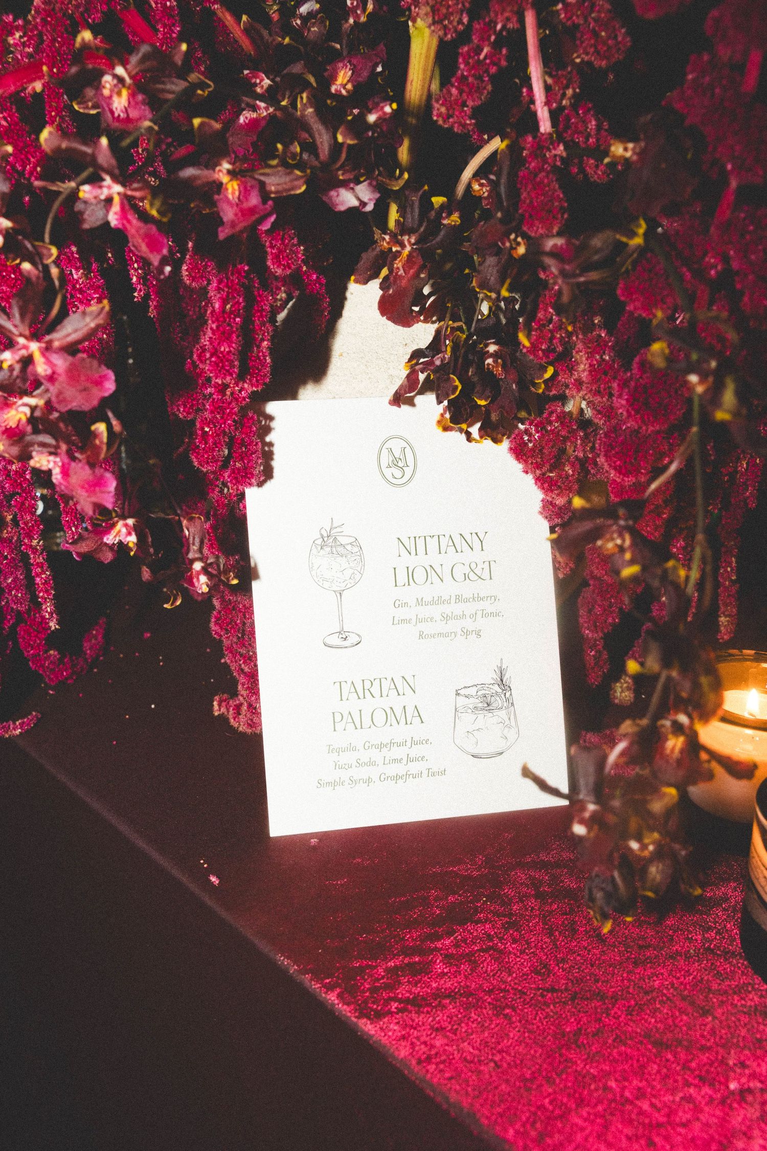 Elegant wedding cocktail menu card surrounded by lush burgundy floral arrangements.