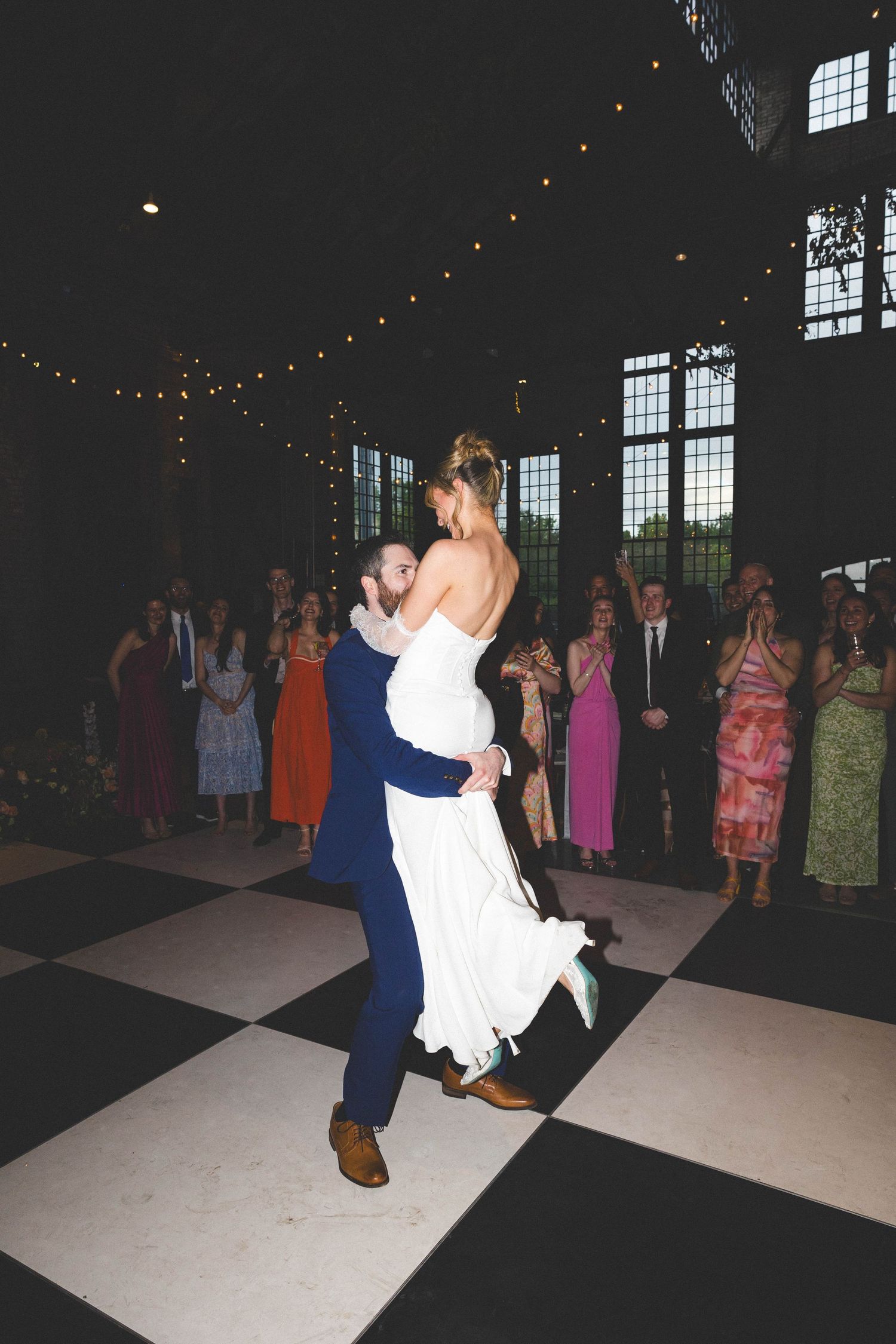 Groom lifting bride in white gown during first dance on checkered floor with guests watching.