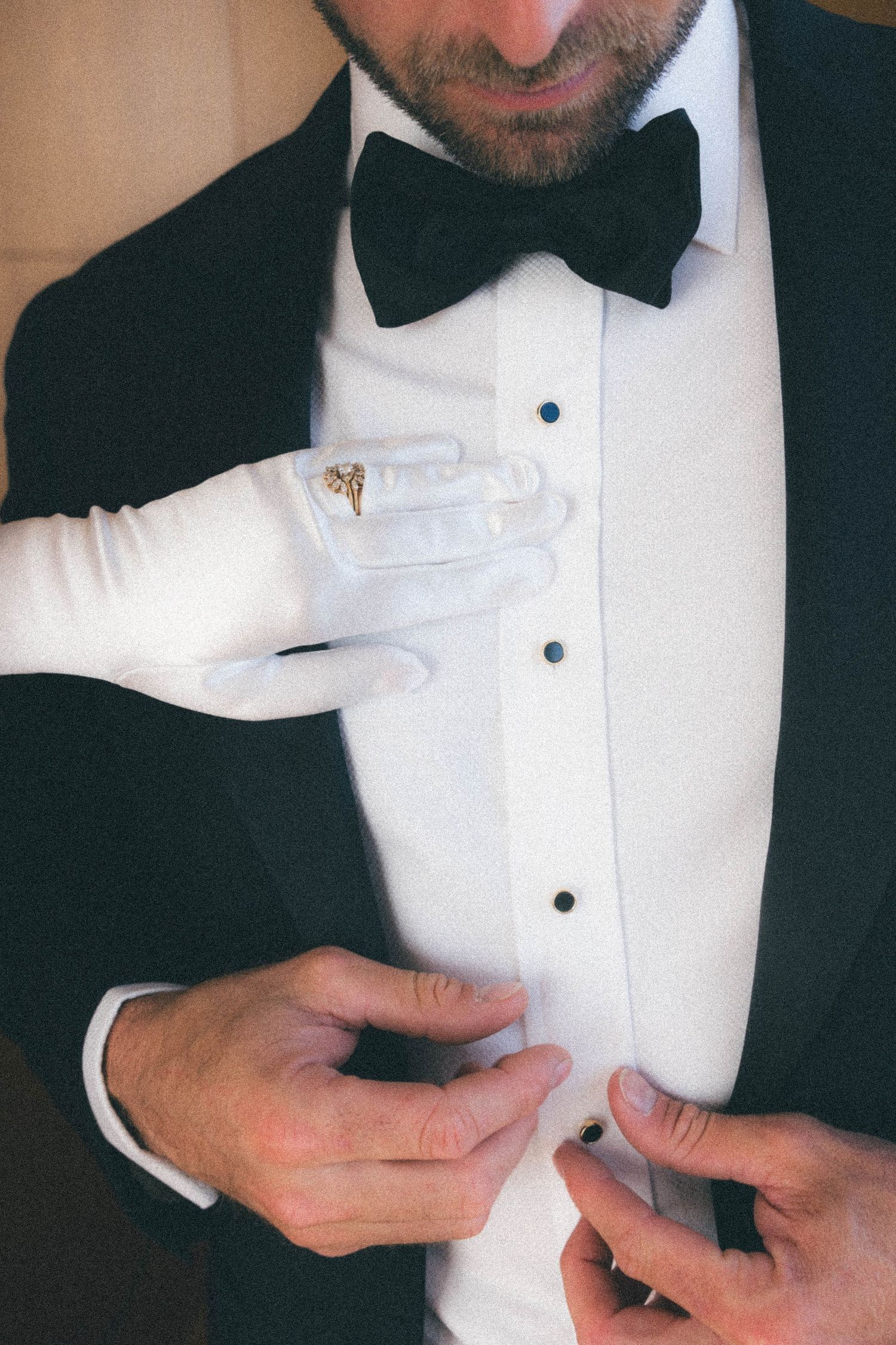 Bride in white gloves pins boutonniere on groom dressed in classic black tuxedo with bow tie.