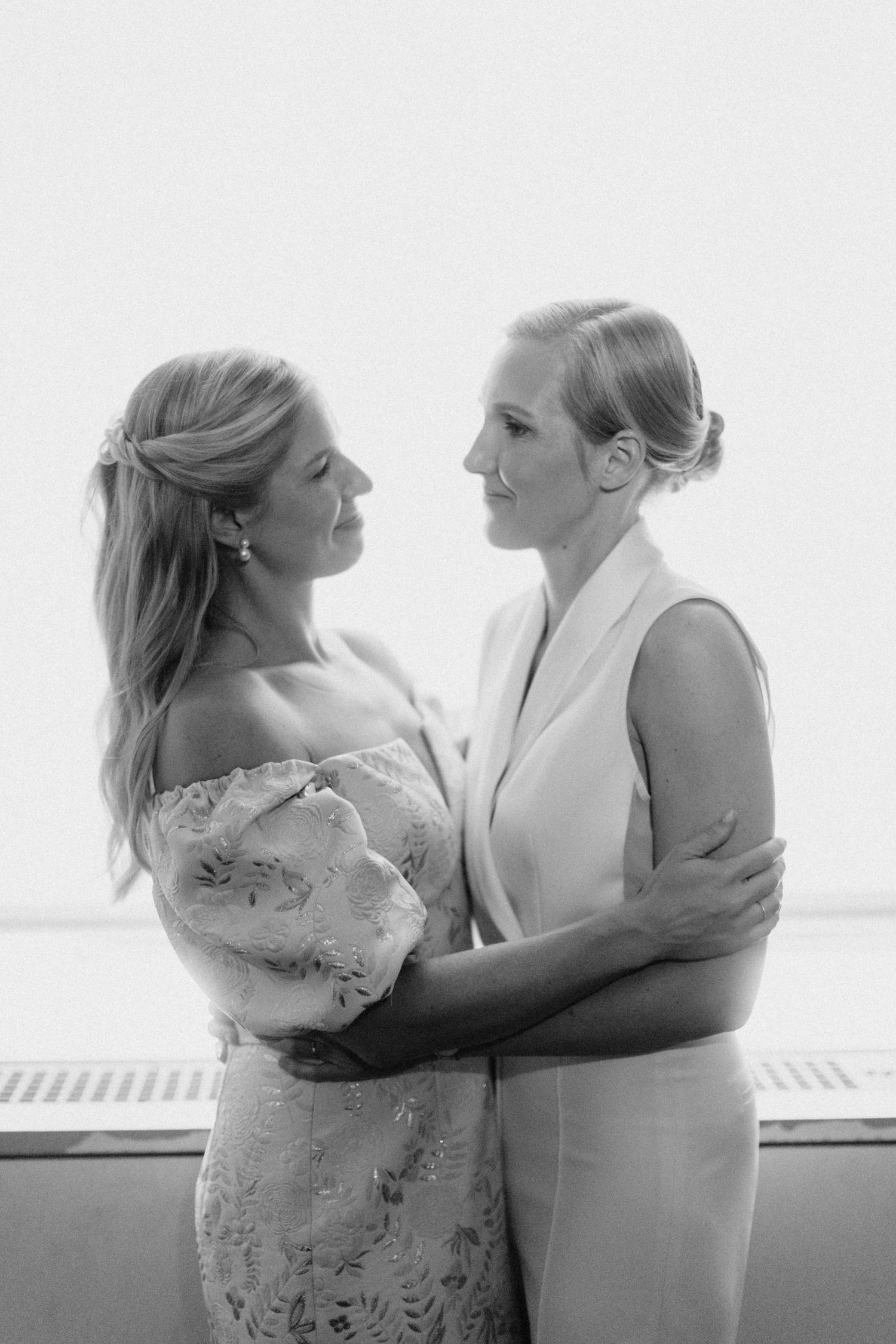 Black and white portrait of two brides embracing and gazing at each other on their wedding day.