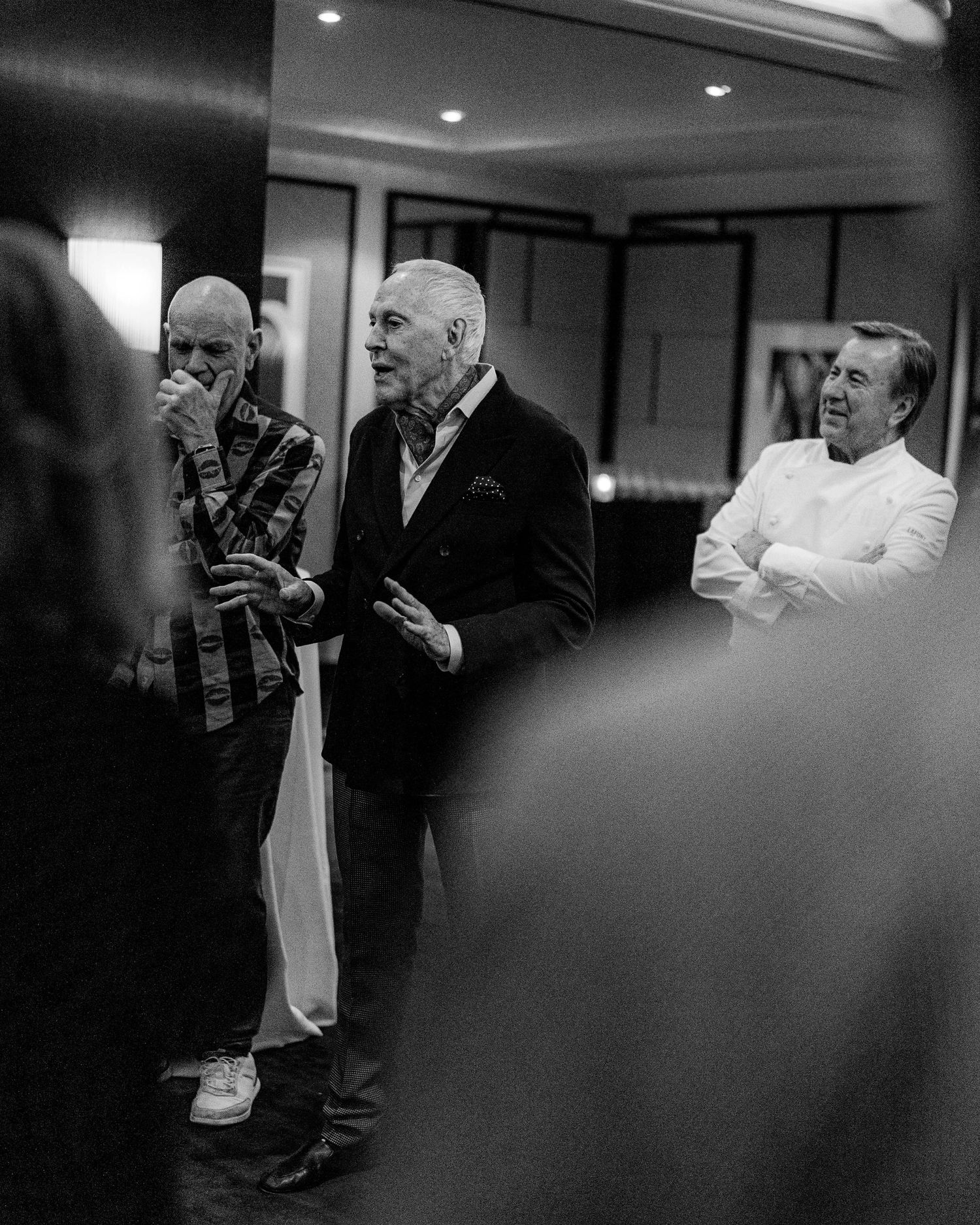 Black and white photo of an older man in a blazer gesturing as he speaks to a room, with two other men standing nearby.