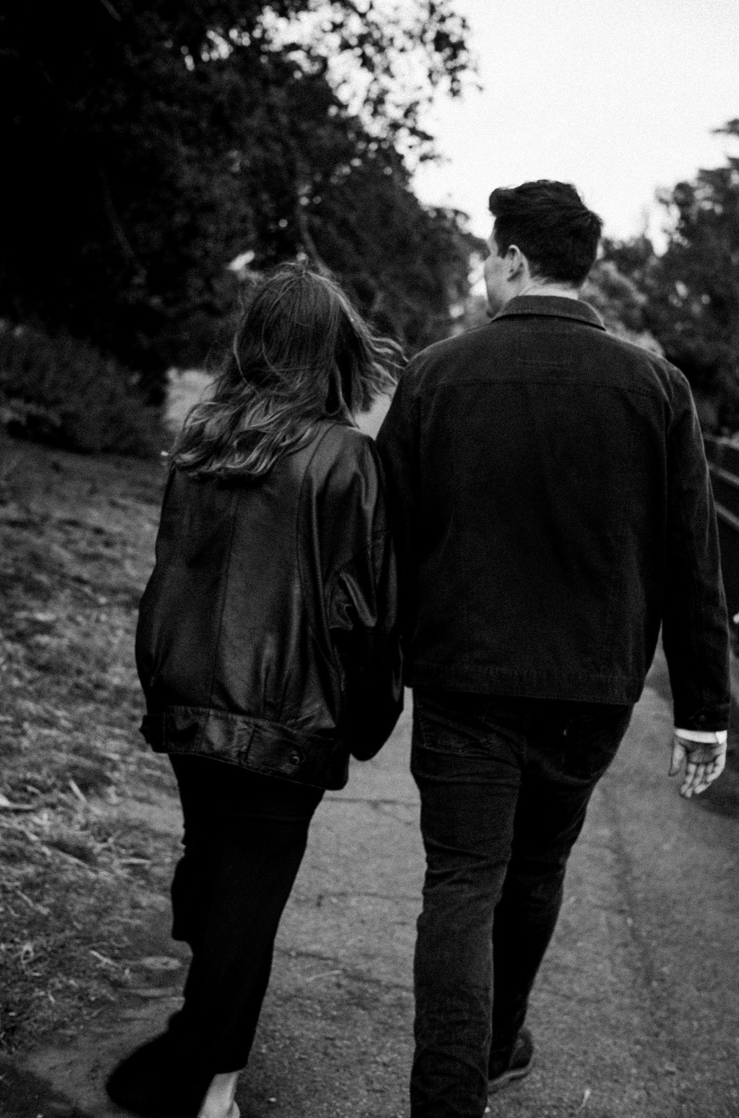 Black and white photo of a couple walking together on a path through a park, viewed from behind.Photography by The Y.K. Studio.