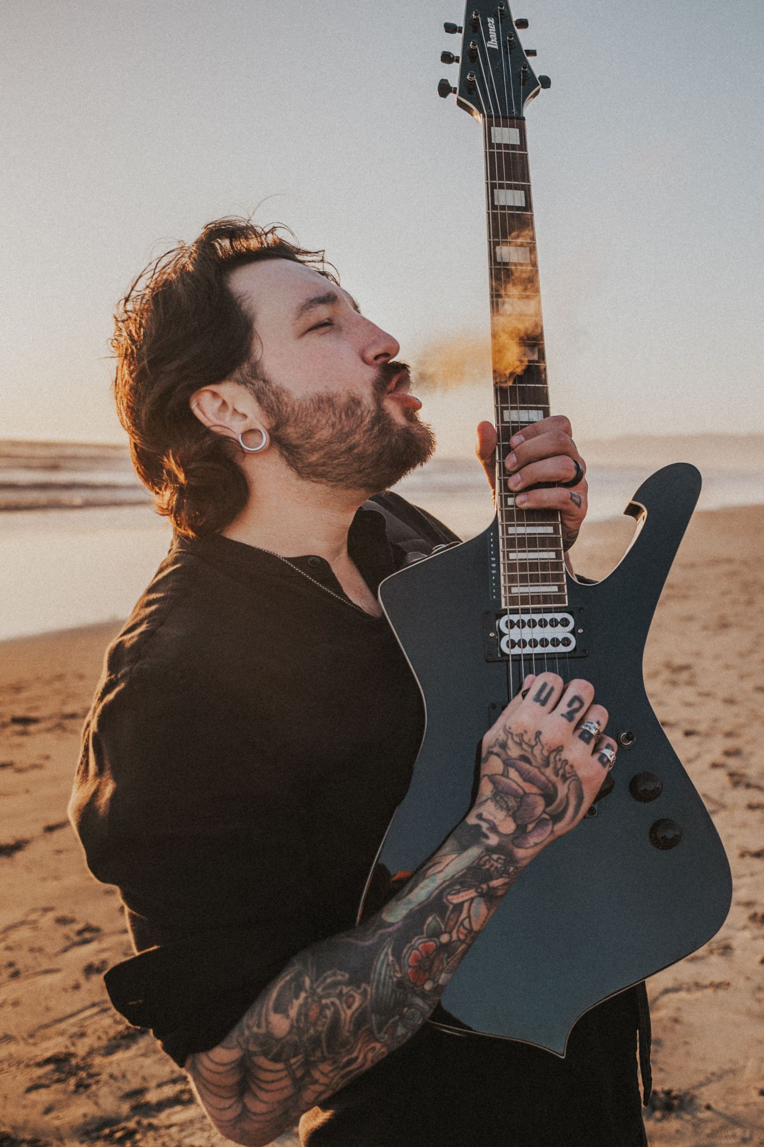 Tattooed man with beard playing dark electric guitar on beach at sunset, wearing black shirt with sleeve tattoos.