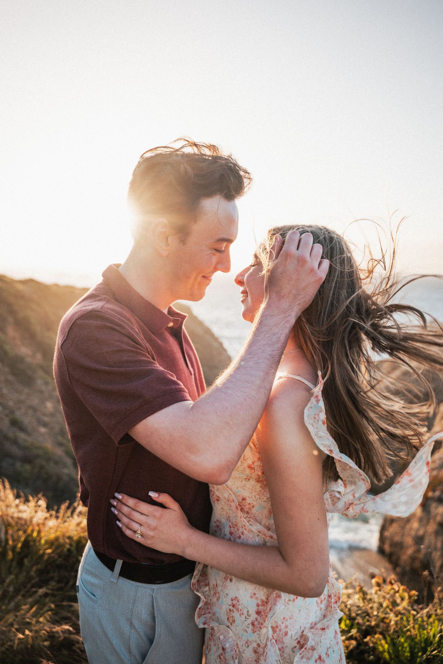 Couple sharing a romantic moment at golden hour on a coastal cliff, man tenderly holding womans hair as wind blows.