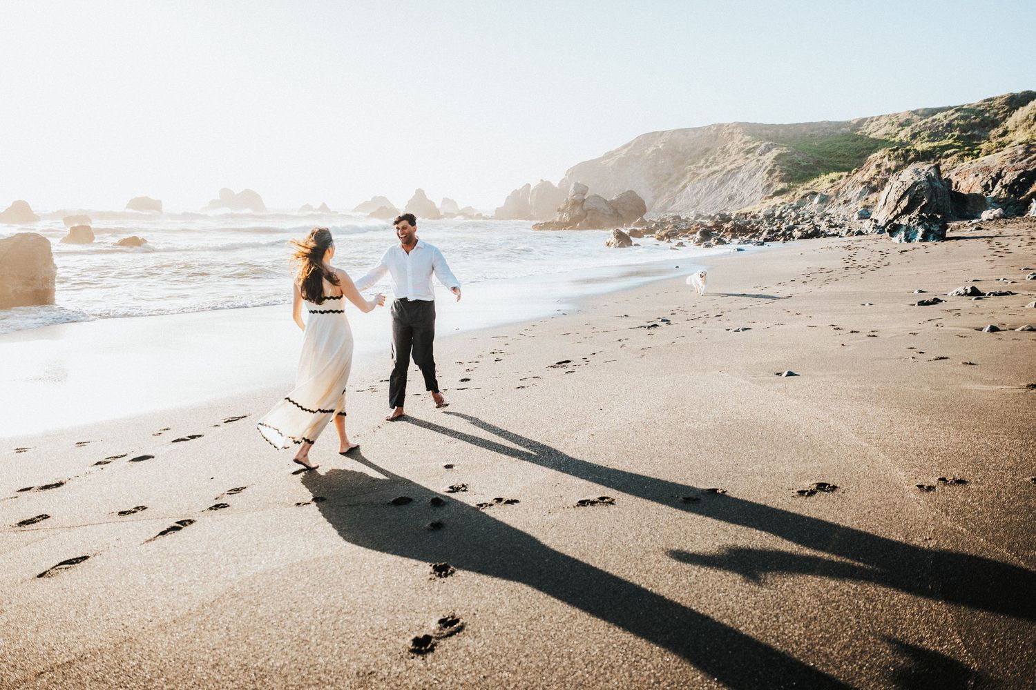 Couple holding hands on a sandy beach with rocky cliffs and ocean waves in golden sunlight.