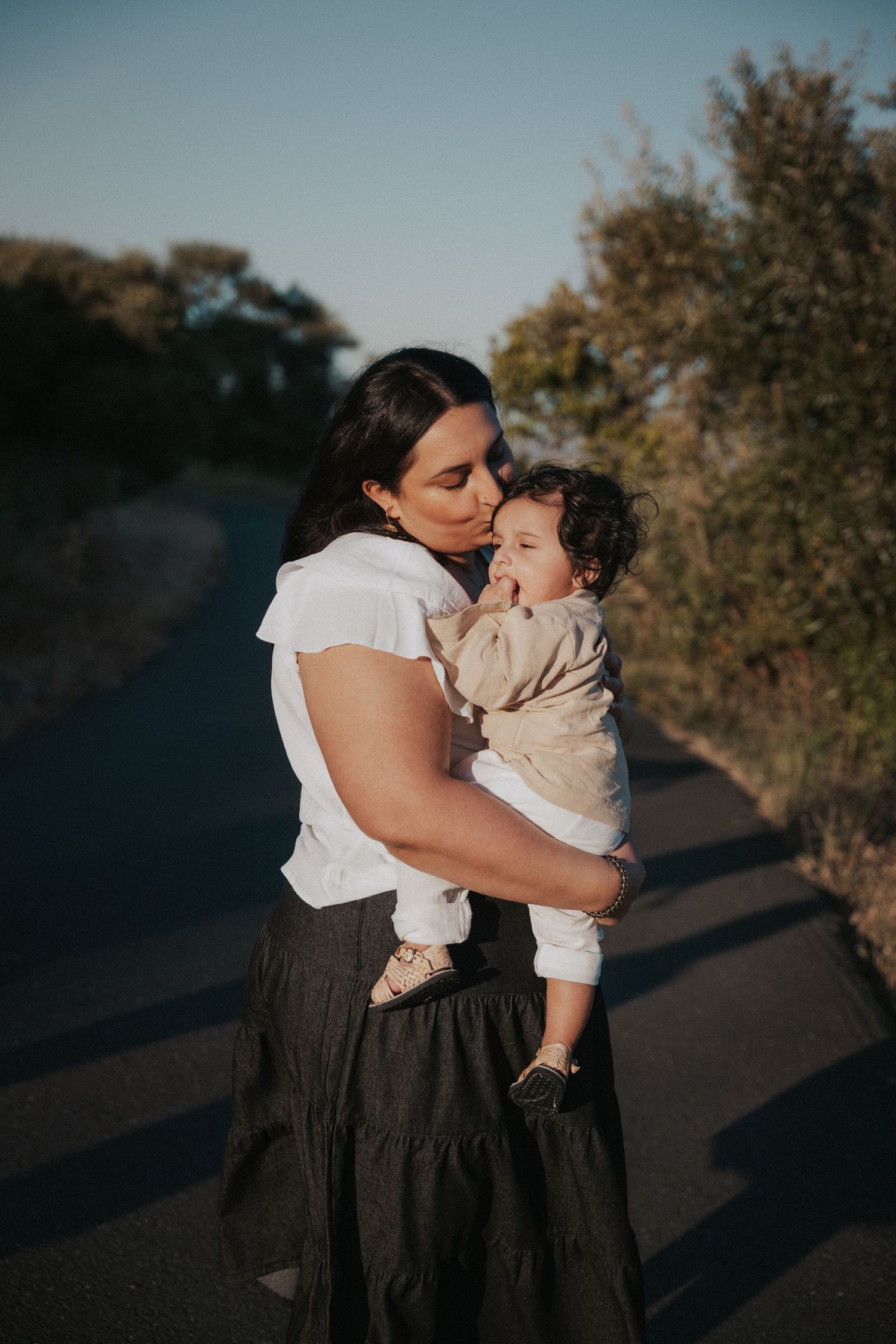 A mother tenderly kisses her baby while holding them on a scenic outdoor path at golden hour.