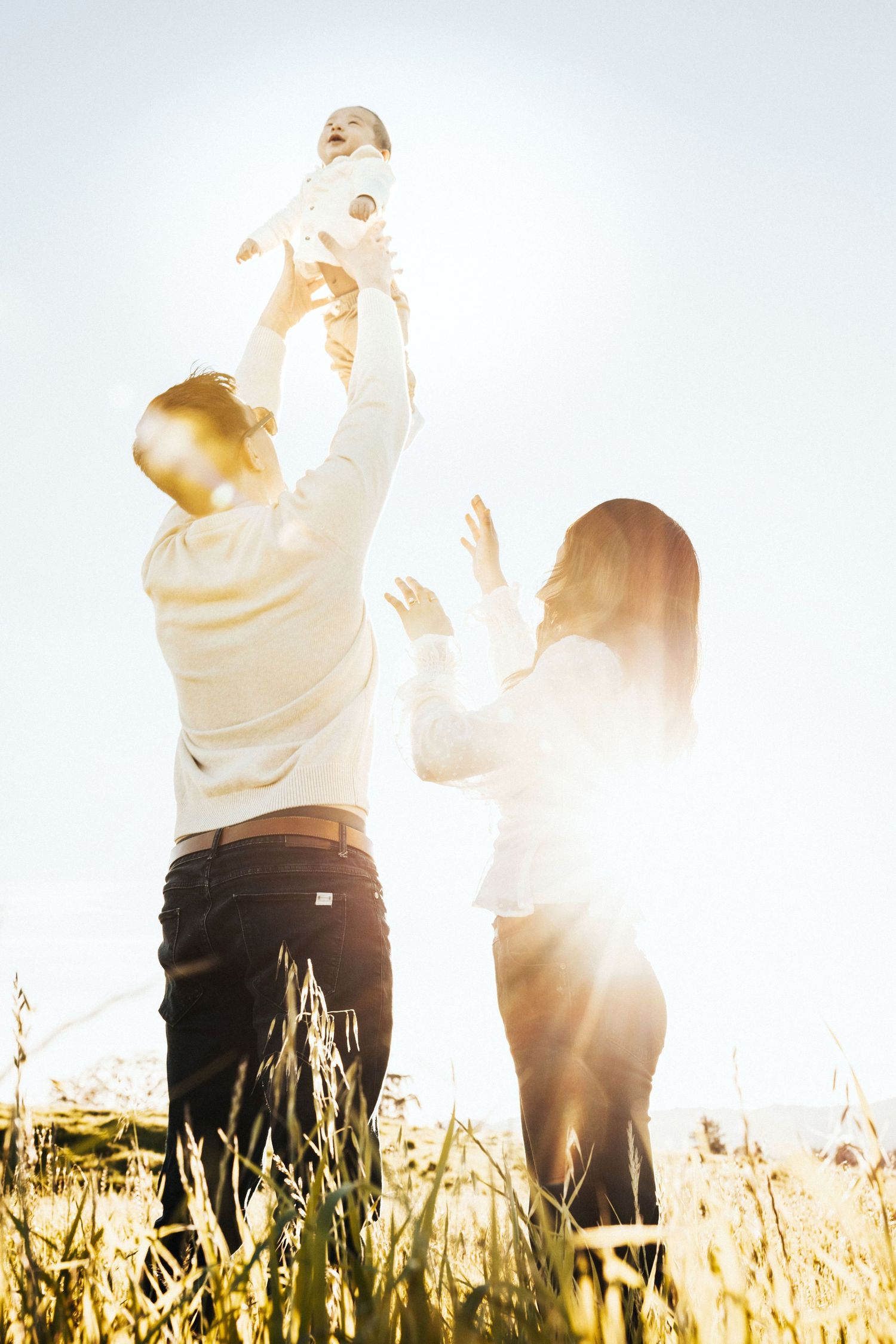 Joyful parents lifting their baby high in the air in a sunlit golden field with bright lens flare.