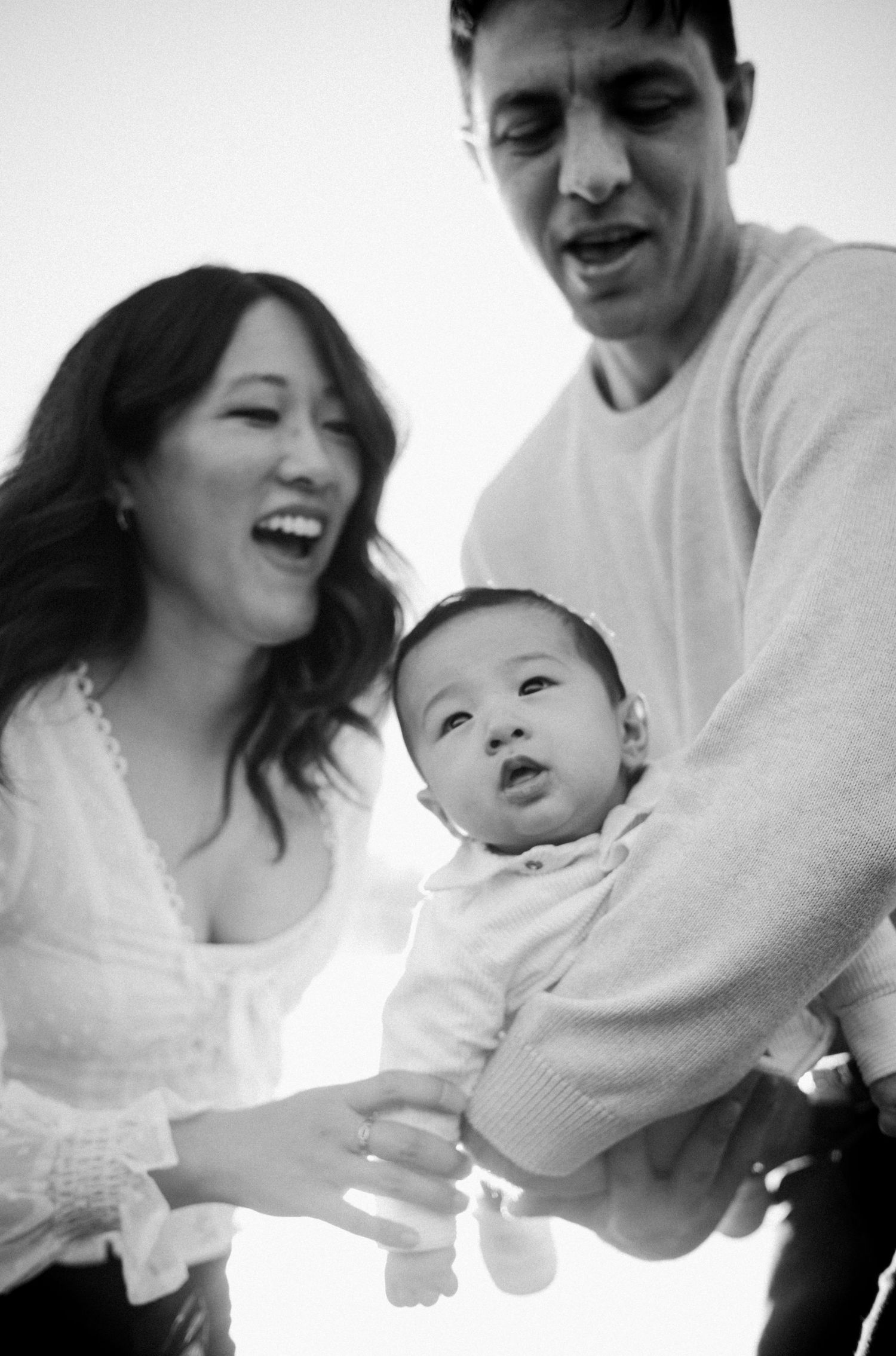 Black and white photo of a smiling Asian woman and man holding their adorable baby, capturing a joyful family moment.