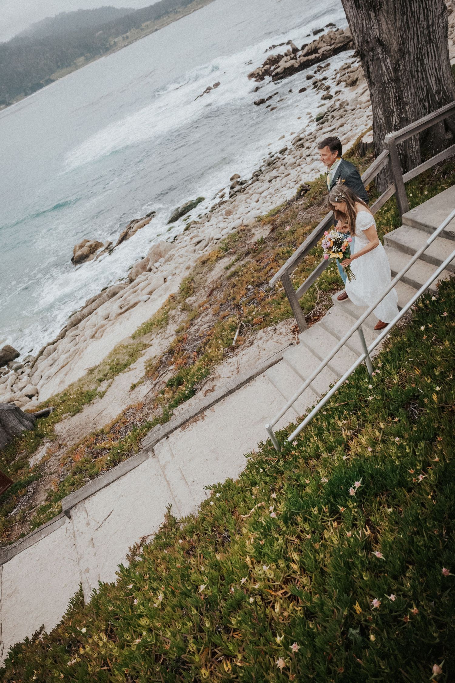 Bride and groom walking down coastal stairs with ocean waves in the background.