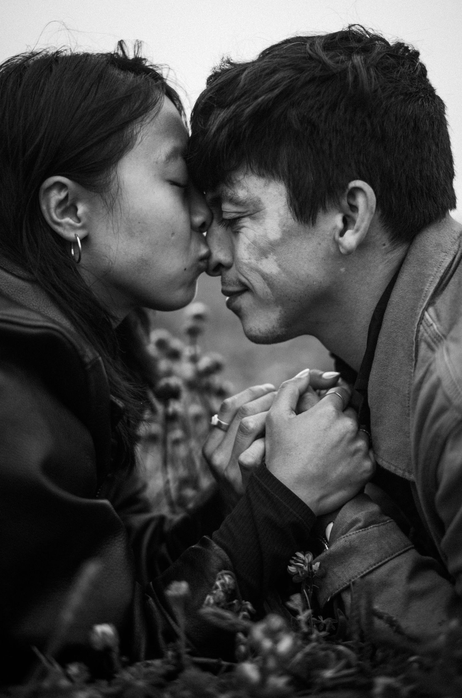 Black and white photo of a couple sharing an intimate nose touch, holding hands tenderly among wildflowers.