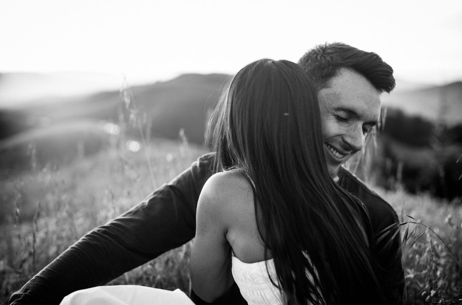 Black and white photo of a smiling couple embracing in a sunlit field with rolling hills in the background.