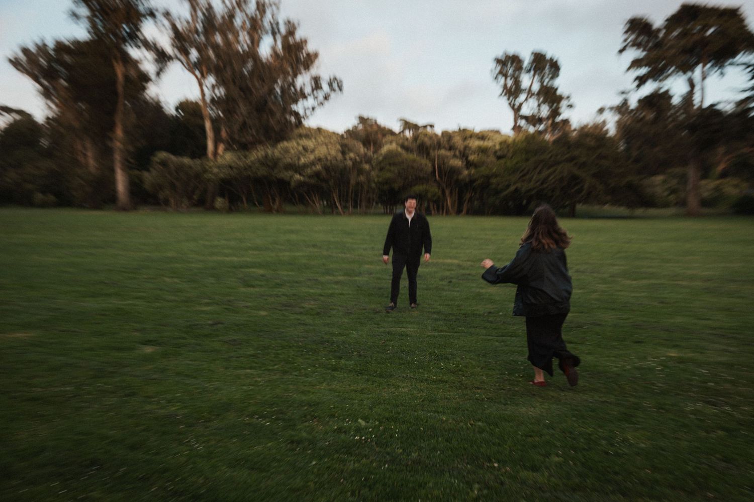 Two people standing in a lush green park field at dusk, surrounded by tall trees in soft evening light.
