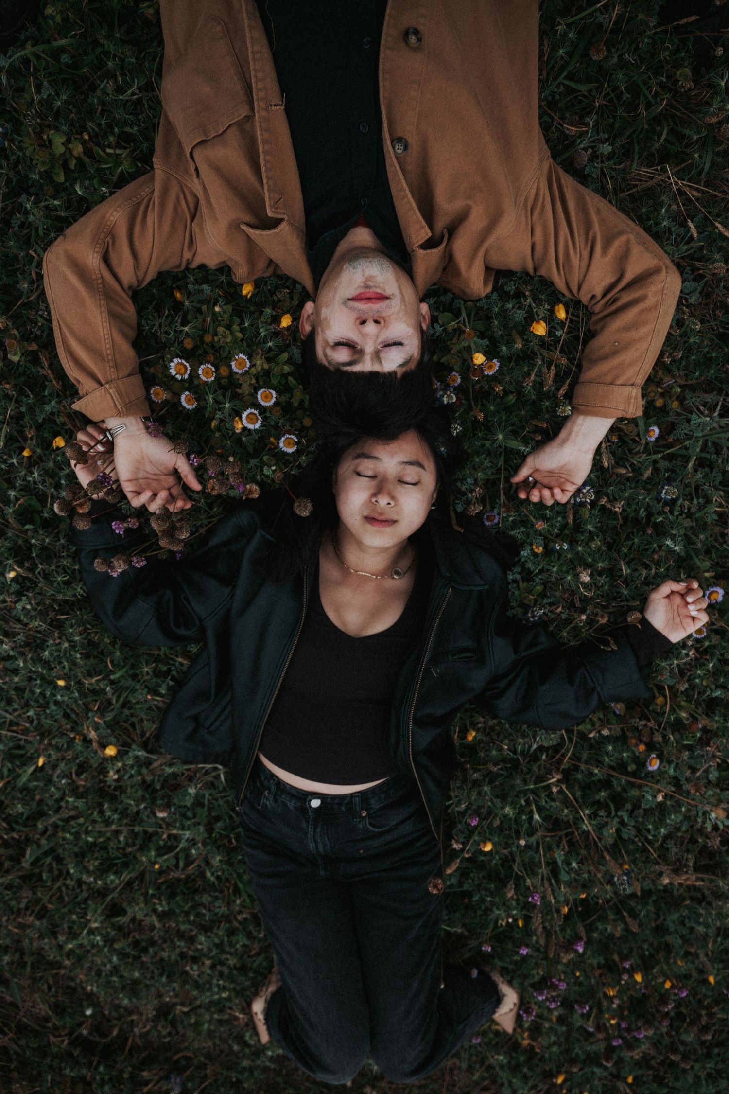 A couple lies on a flower-covered grass field with eyes closed, wearing brown and black jackets.