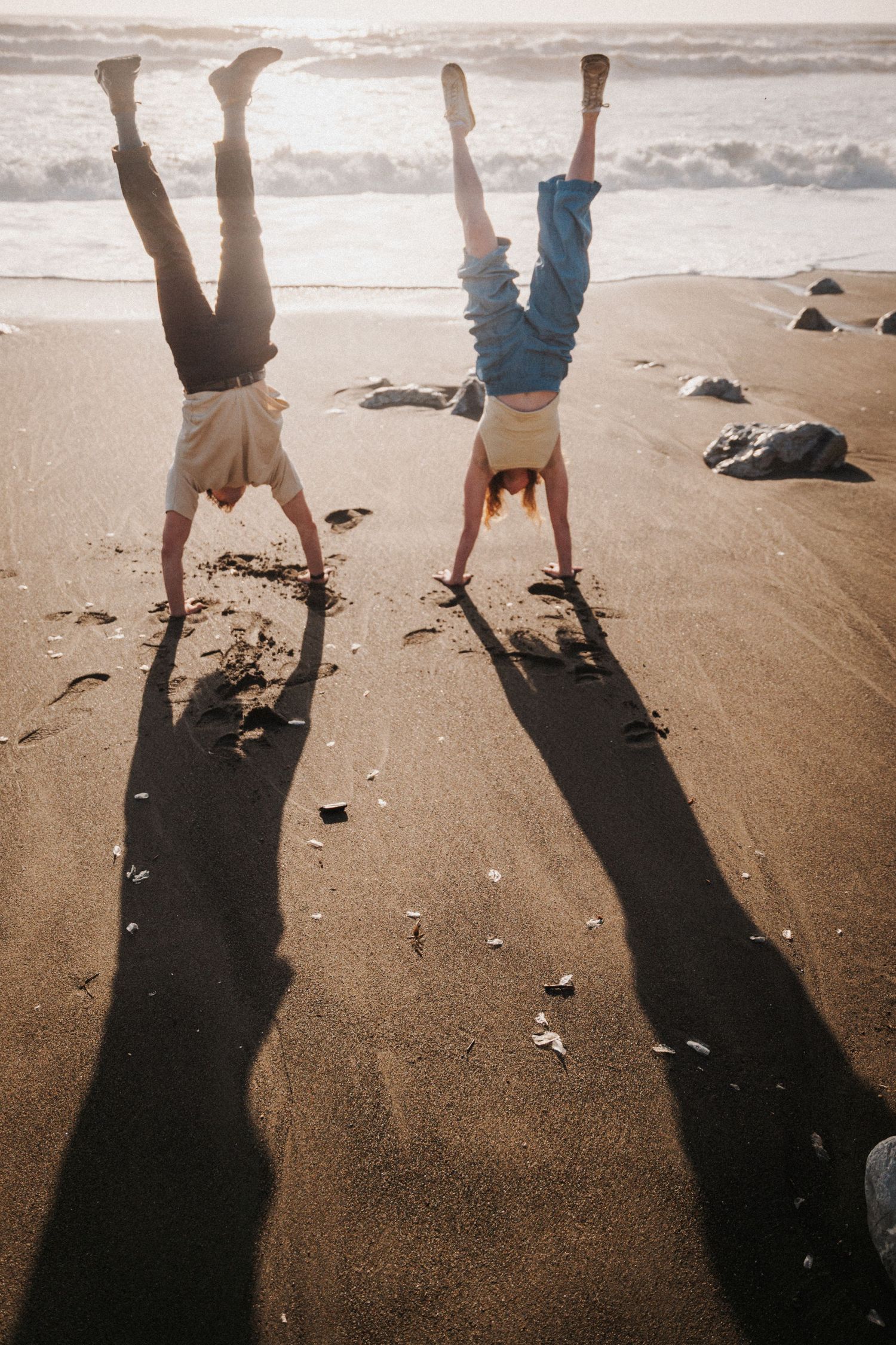 Two people doing handstands on a sandy beach at sunset, casting long shadows on the shore with ocean waves in the background.