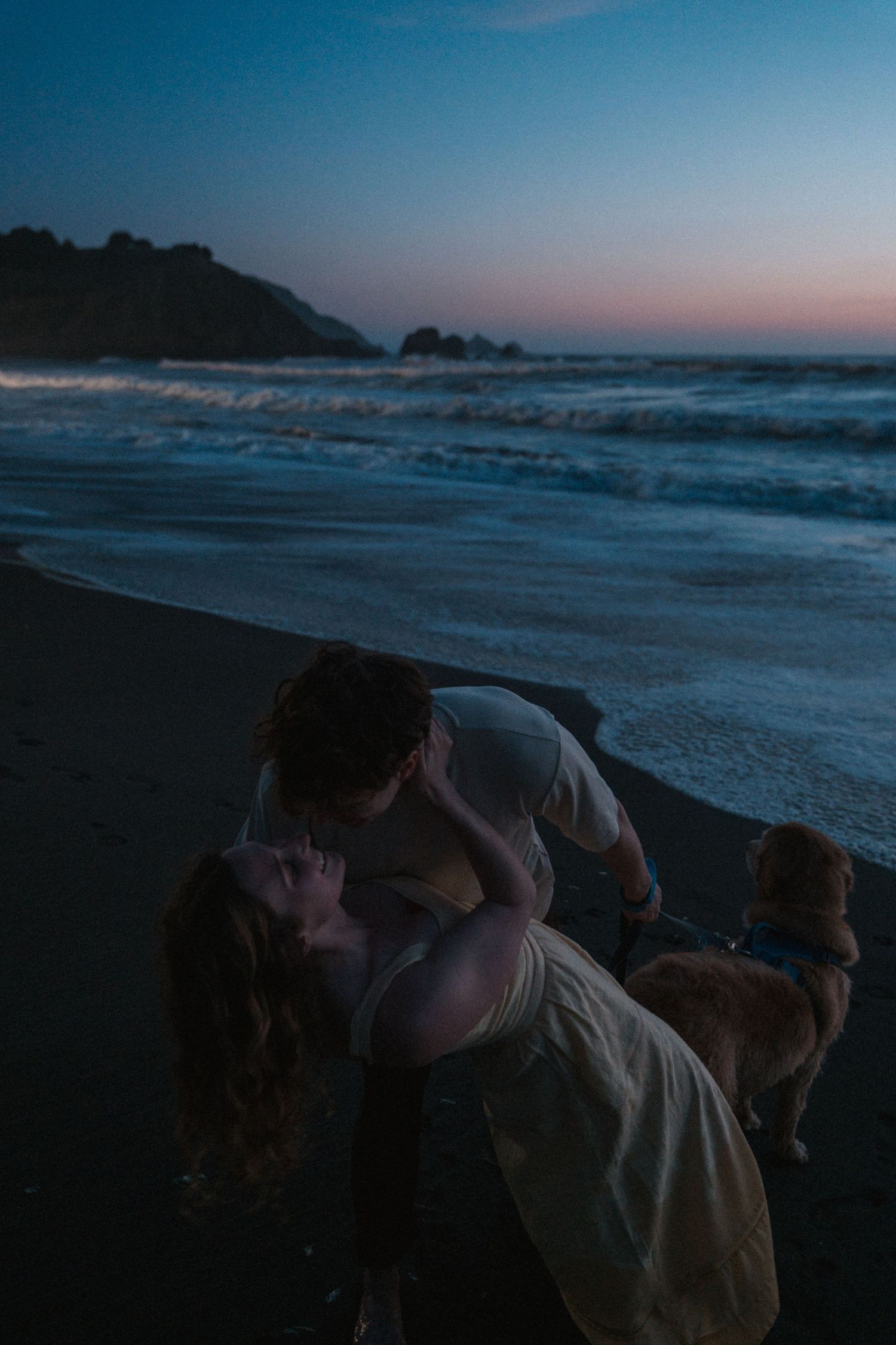 Couple sharing a romantic dip kiss on a dark beach at twilight with waves and a dog nearby. Photography by The Y.K. Studio.