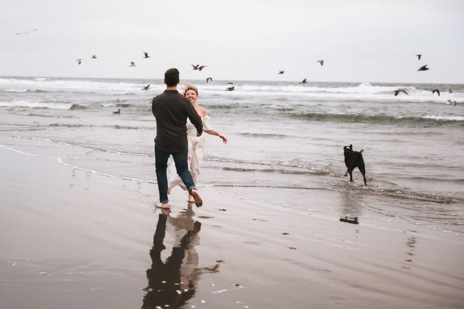 Couple running playfully on a beach with a black dog and birds flying over ocean waves on a cloudy day.