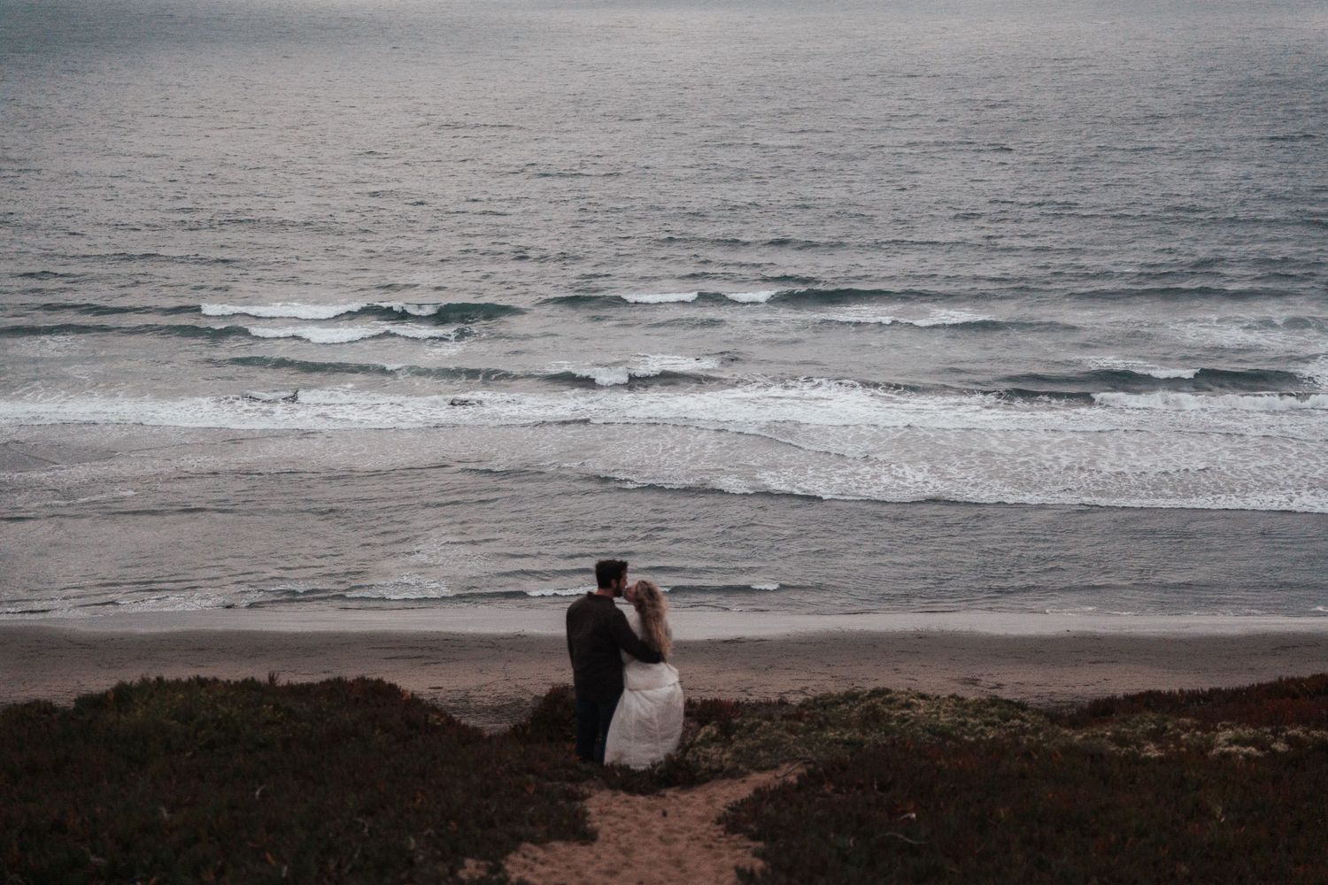 Couple in wedding attire embracing on a cliffside overlooking ocean waves at a romantic beach elopement.