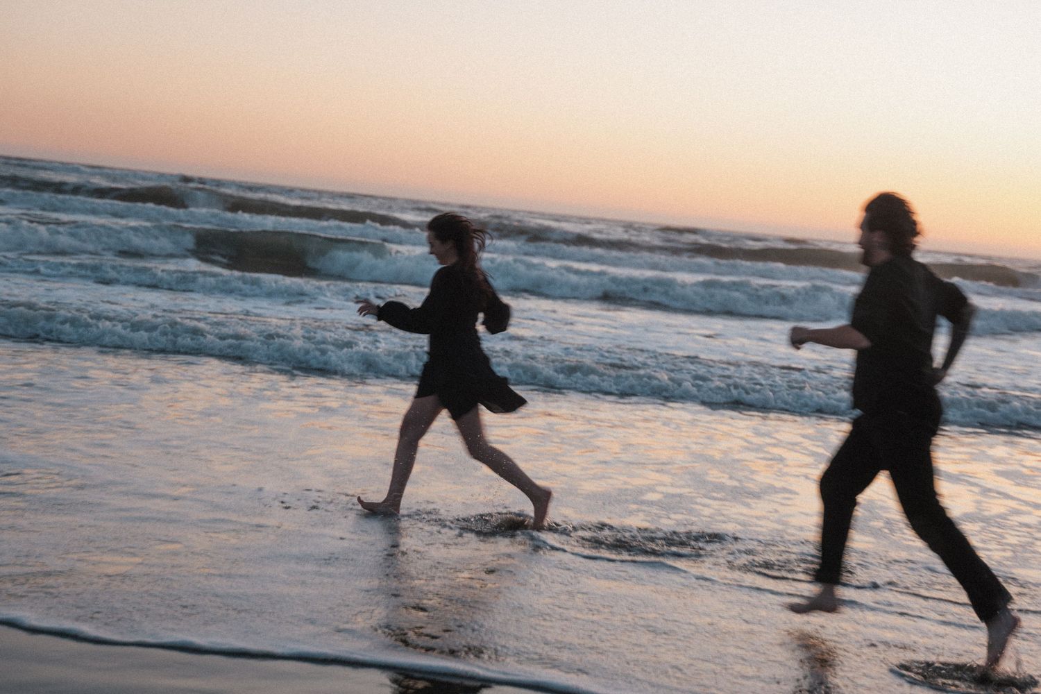 Silhouetted couple running barefoot along ocean shoreline at sunset on sandy beach.