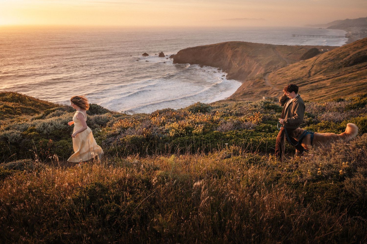 Couple walking on golden coastal cliffs at sunset with ocean views.