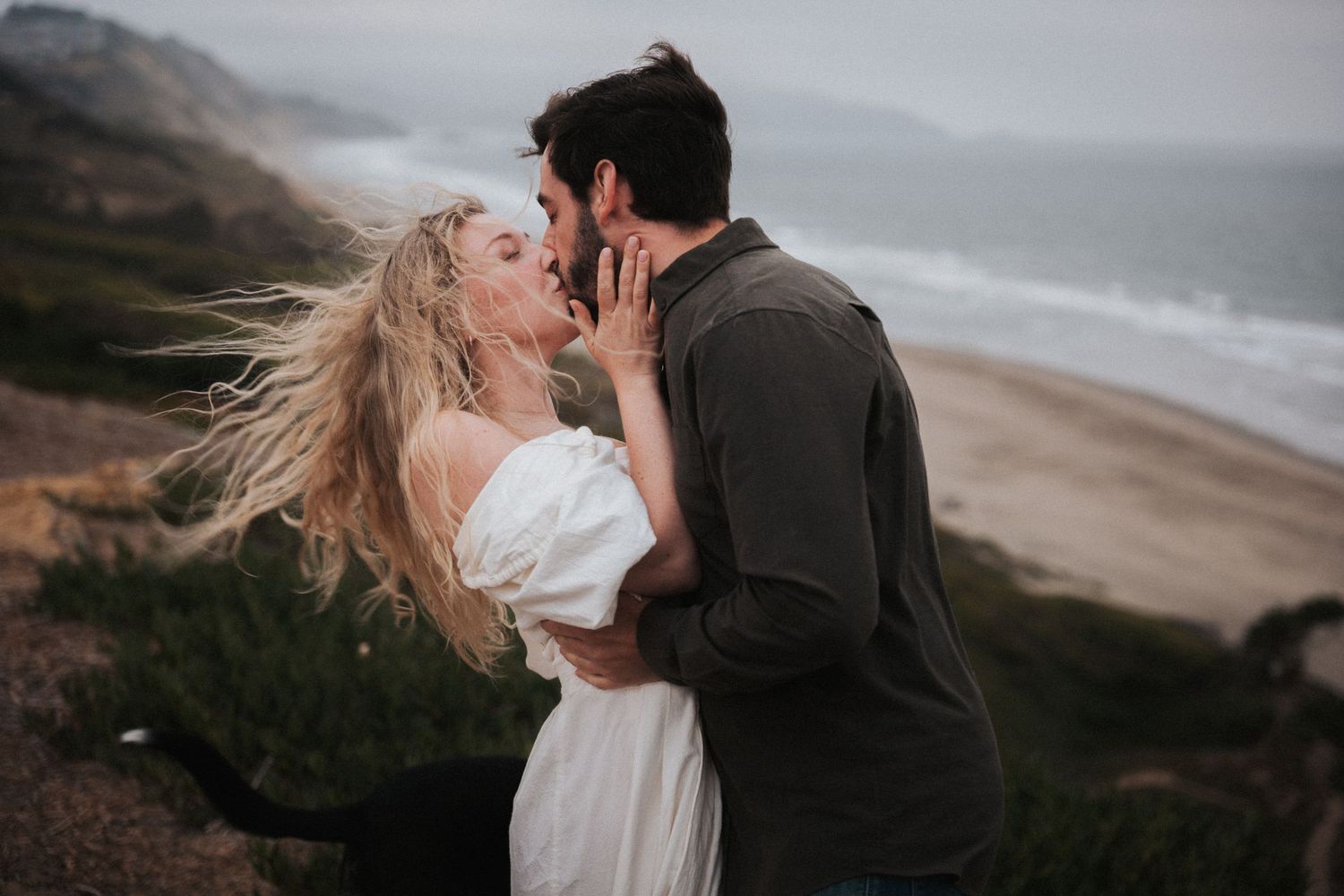 Couple sharing a romantic kiss on a cliffside beach, woman in white dress with wind-blown hair, moody coastal backdrop.