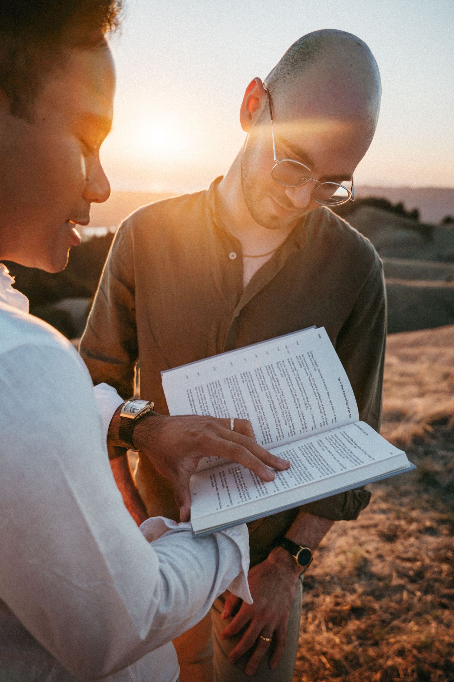 Two people reading a book together outdoors at golden sunset, man with glasses smiling while woman points at page.
