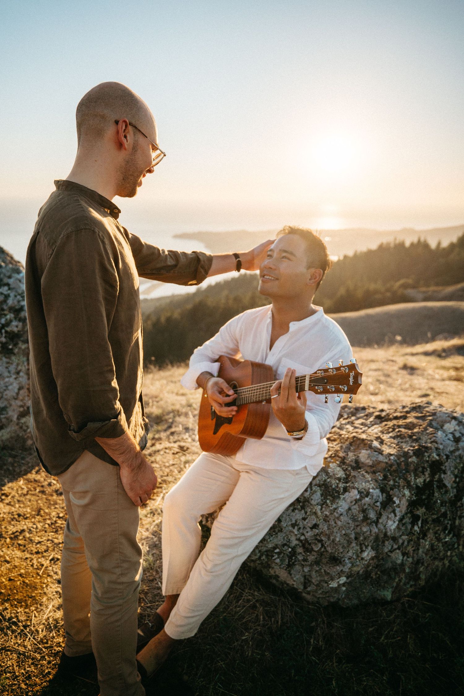 Two men share a romantic sunset moment on a hilltop, one playing acoustic guitar while the other gently touches his head.