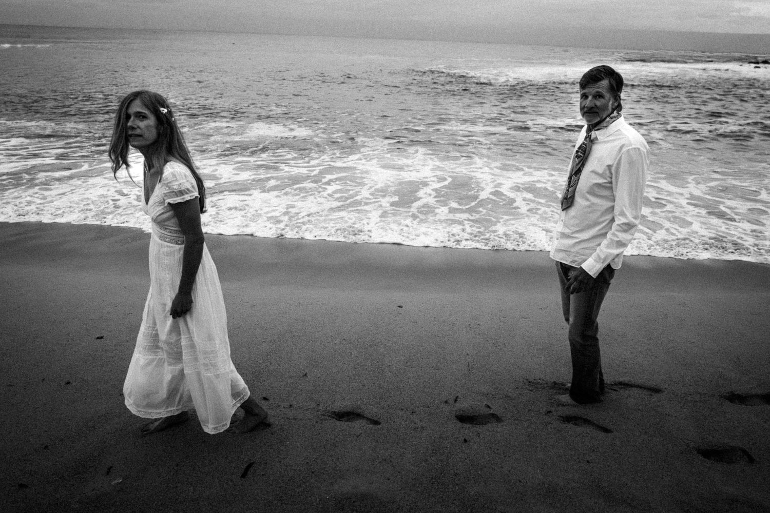 Black and white beach photo of a couple standing apart on wet sand with ocean waves behind them.Photography by The Y.K. Studio.