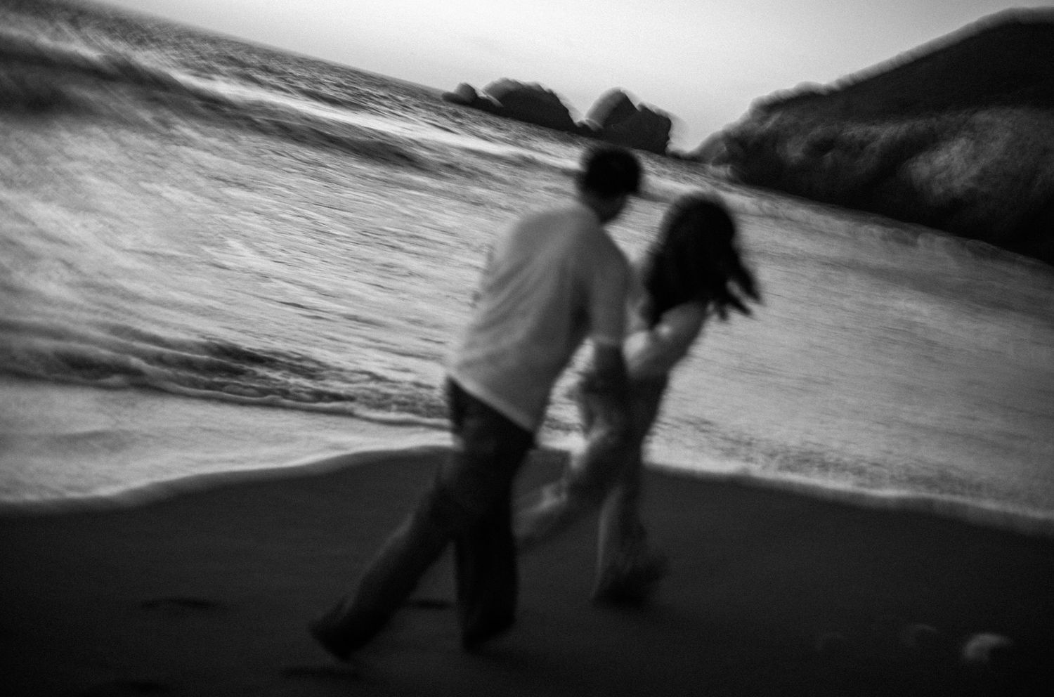 Black and white photo of a couple dancing playfully on a beach with ocean waves in the background.