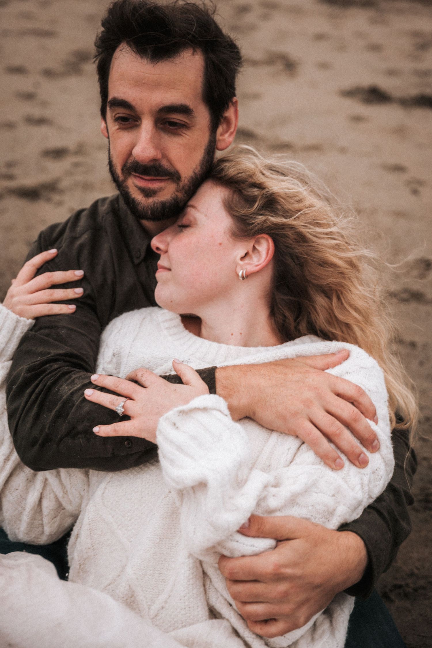 Bearded man tenderly embracing blonde woman in white sweater while sitting on sandy beach.