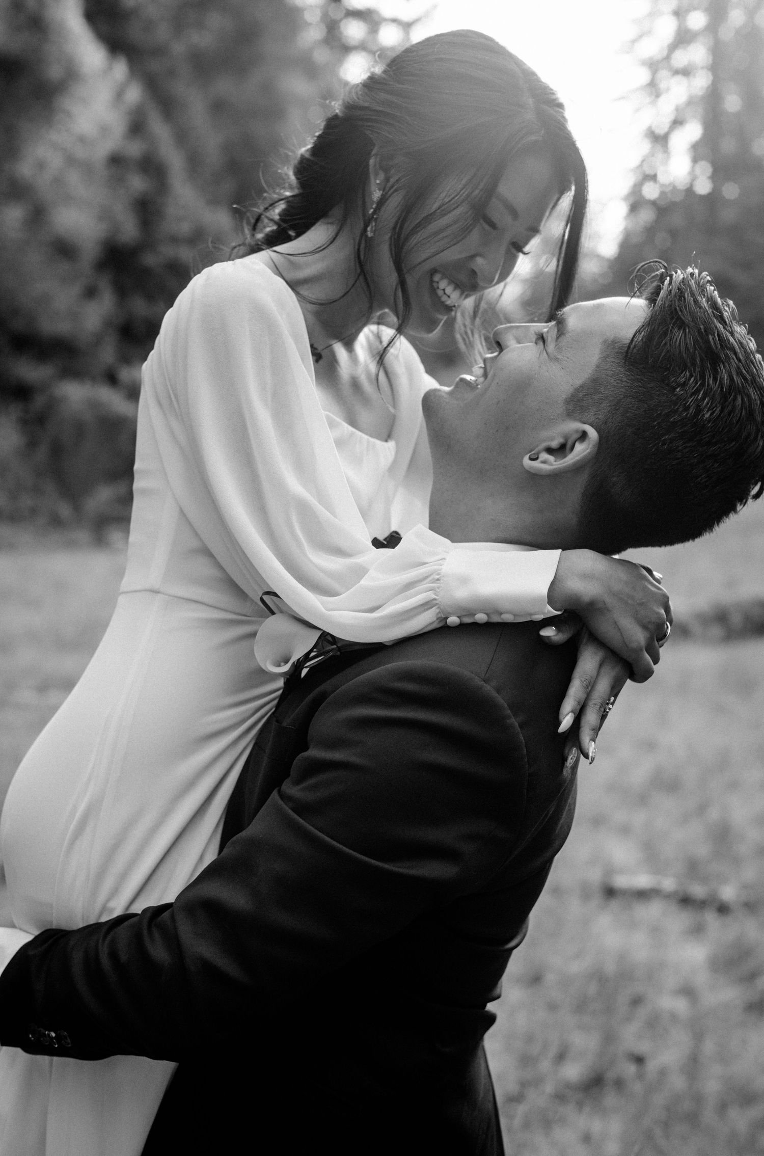 Black and white photo of groom lifting laughing bride in white dress as they share a joyful romantic moment outdoors.