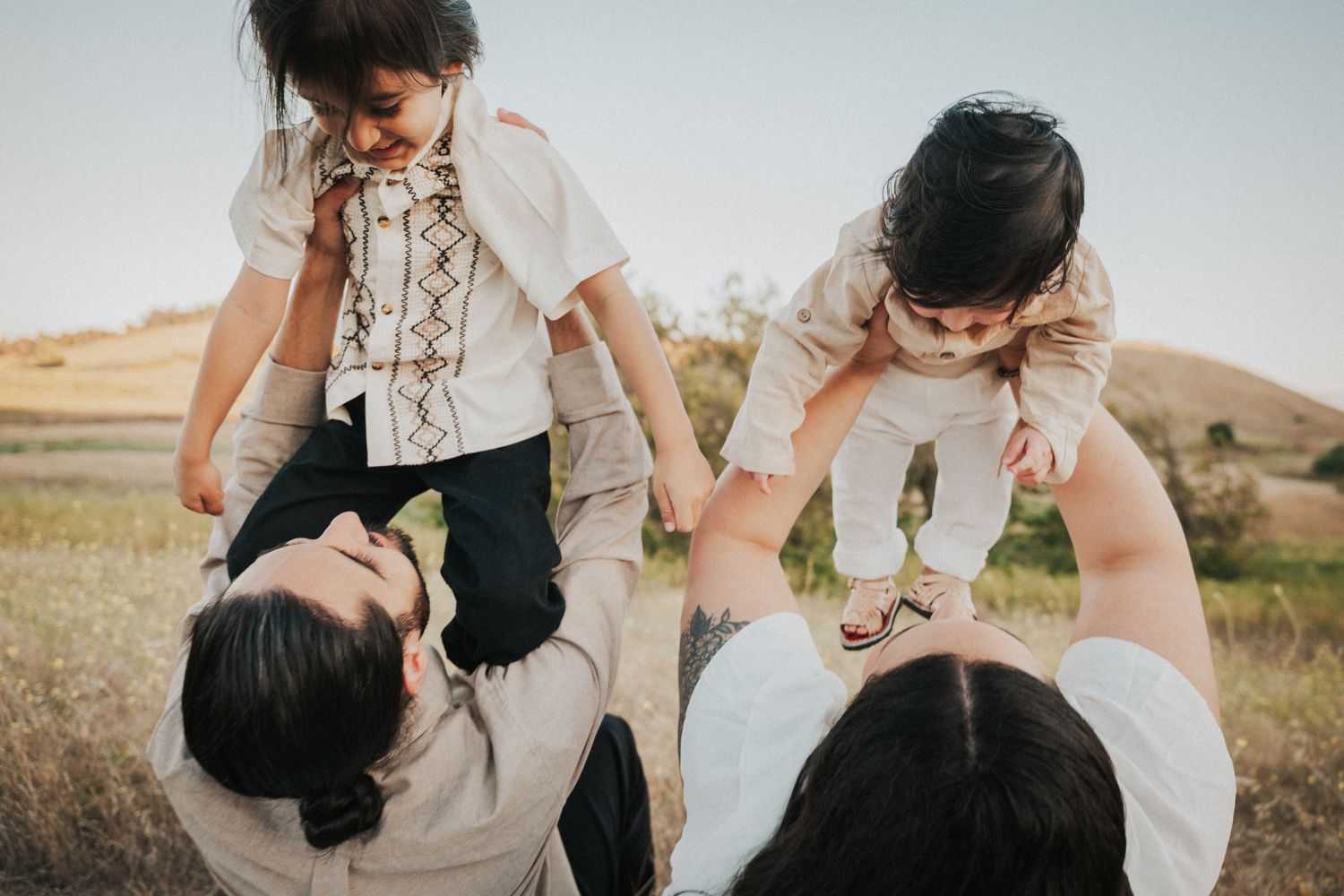 Two parents playfully lifting their children in a sunny field, capturing a joyful family moment outdoors.