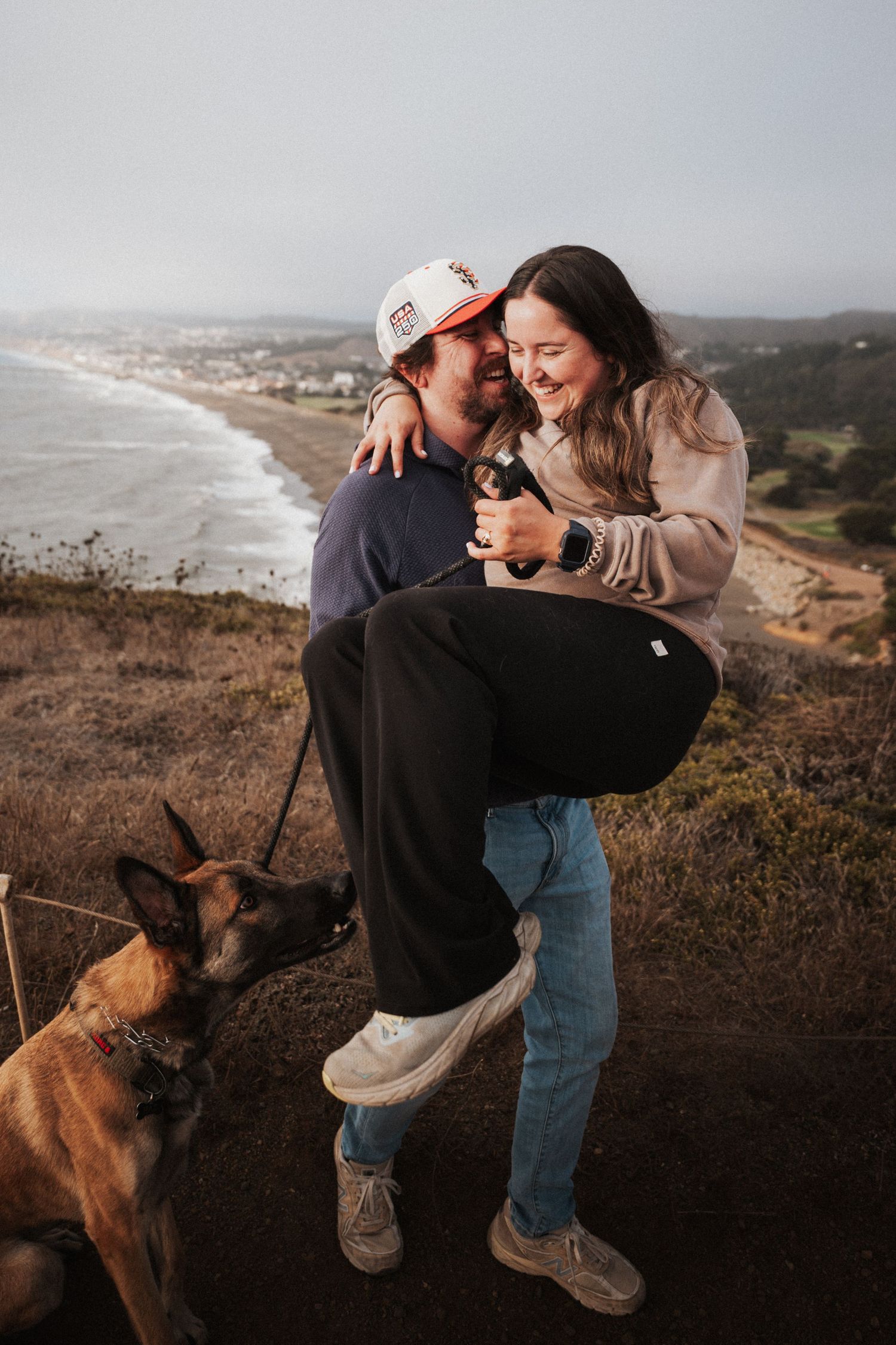 Couple laughing on coastal bluff, man lifting woman, Belgian Malinois dog nearby, ocean and foggy shoreline in background.