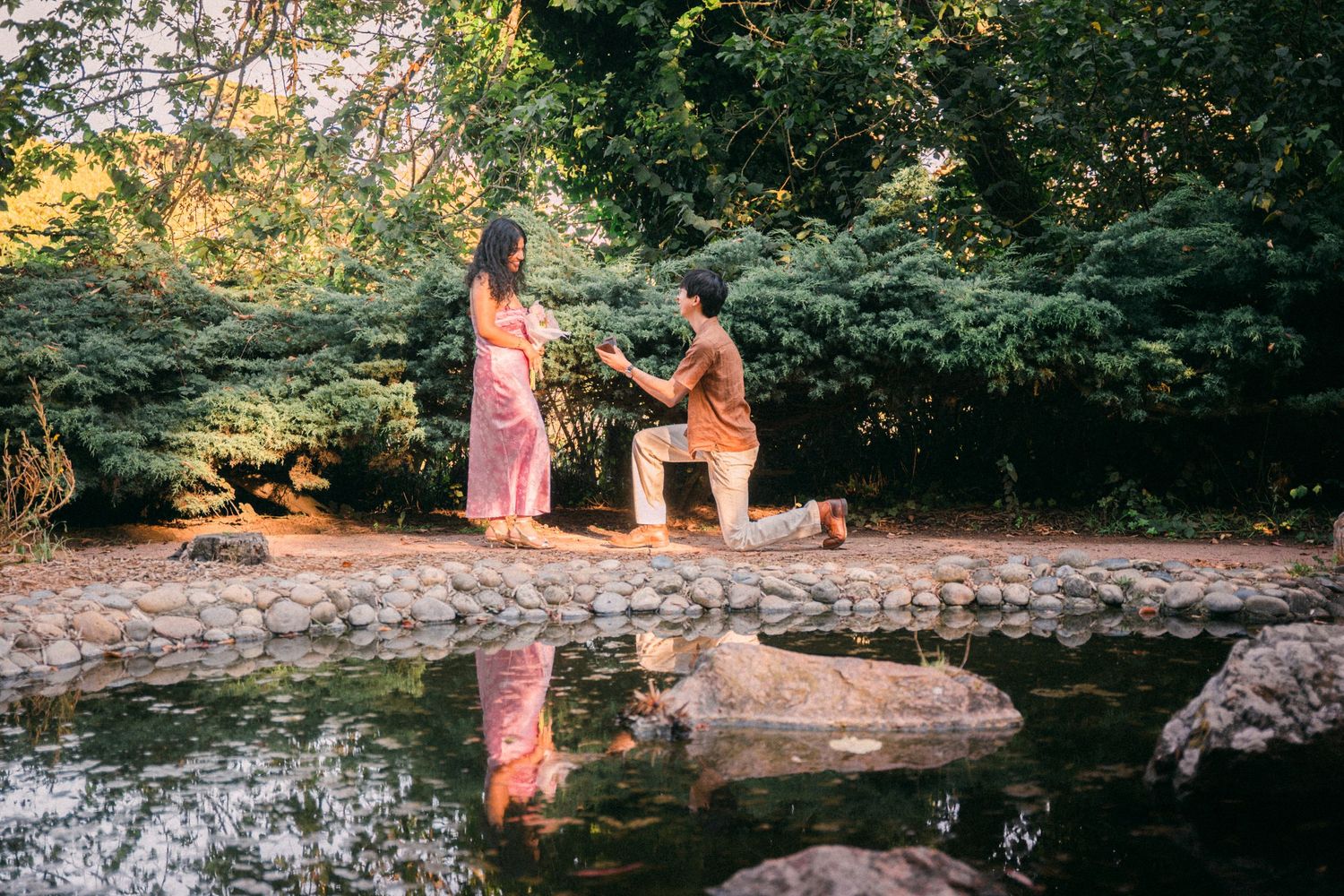 Romantic marriage proposal by a reflective pond in a lush green botanical garden setting.
