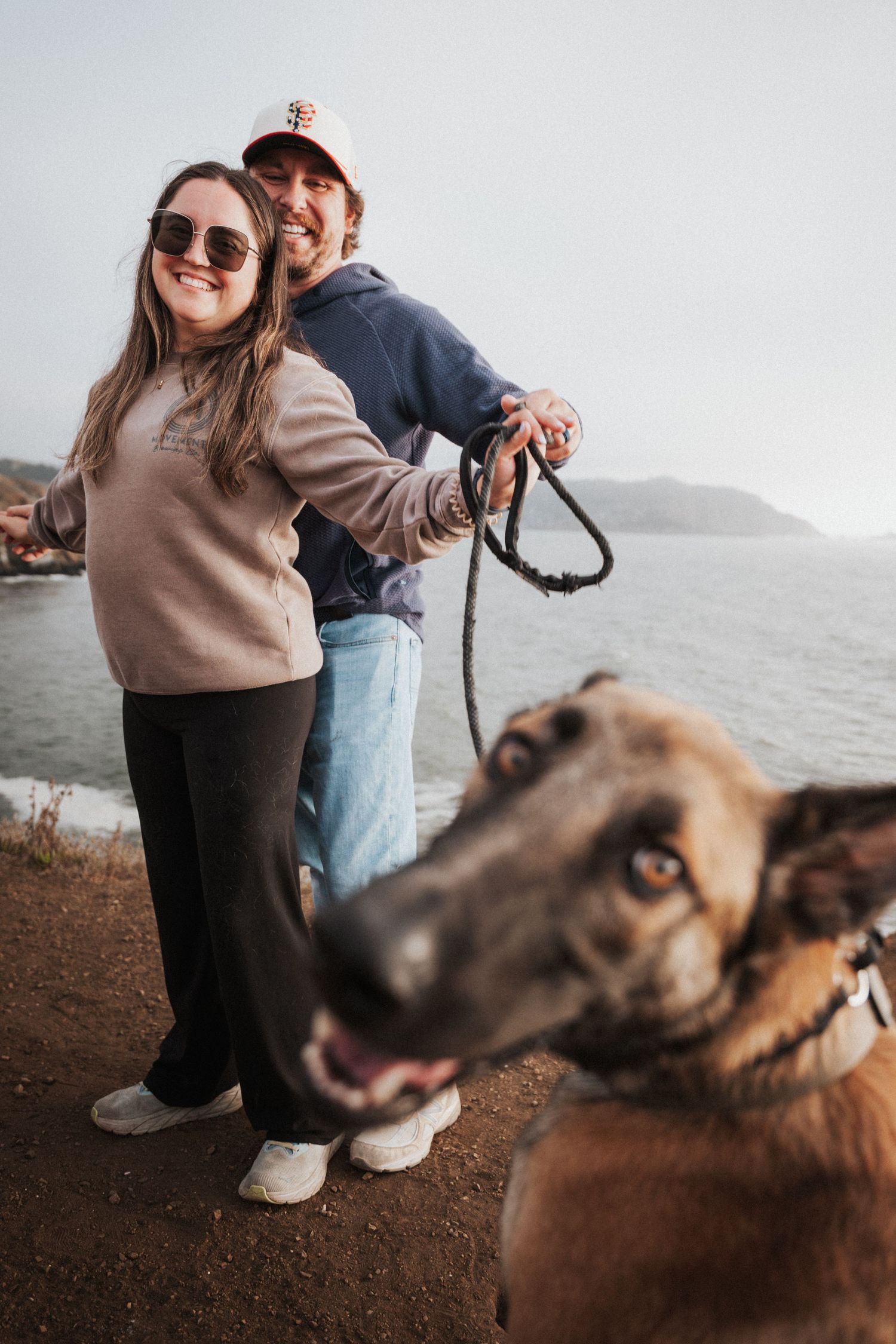 Couple smiling outdoors near ocean with German Shepherd dog on leash in foreground, wearing casual hoodies near coastal cliffs.