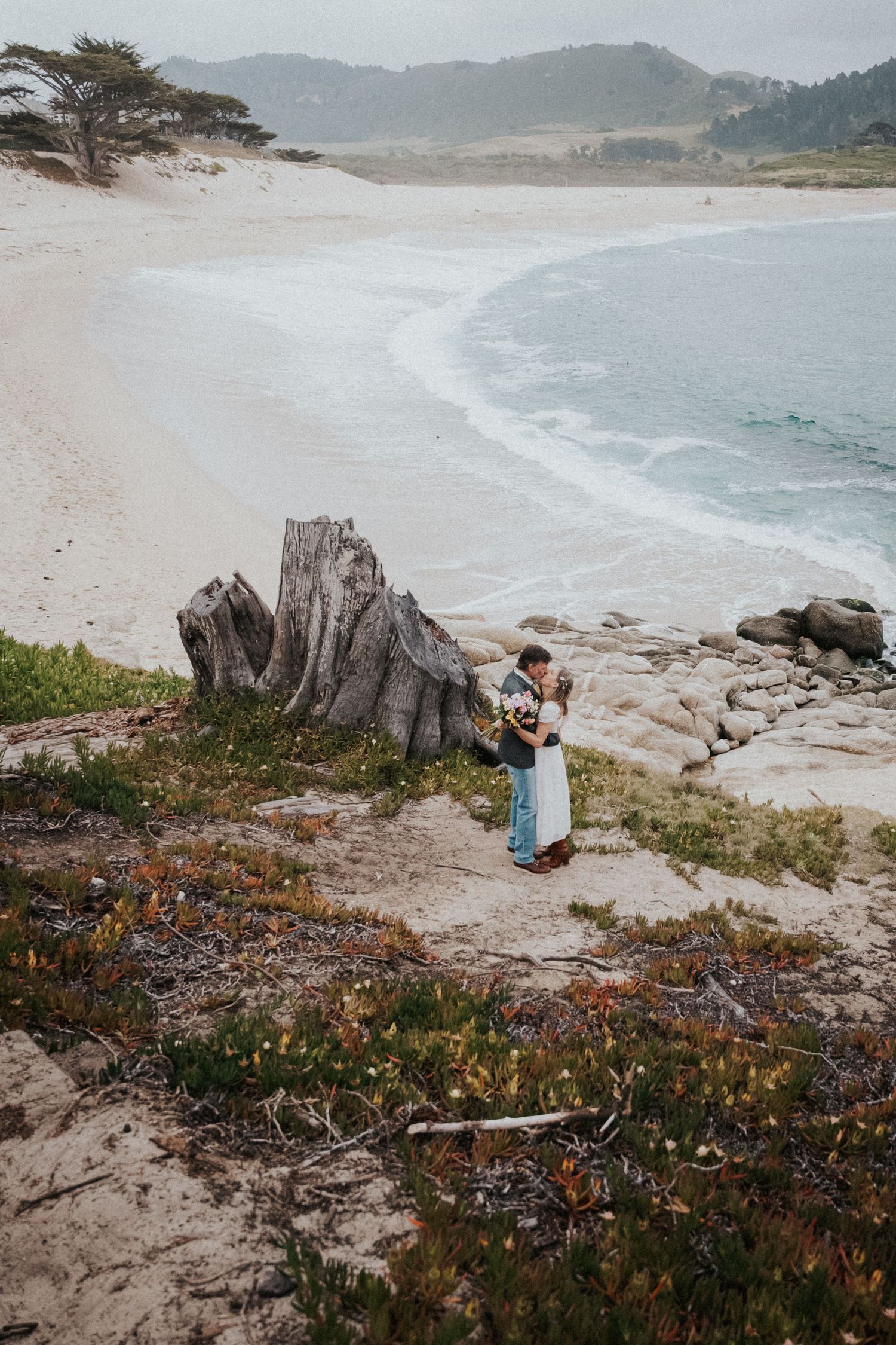 Couple embracing near driftwood stump on rocky Carmel Beach coastline with white sand and blue ocean waves.