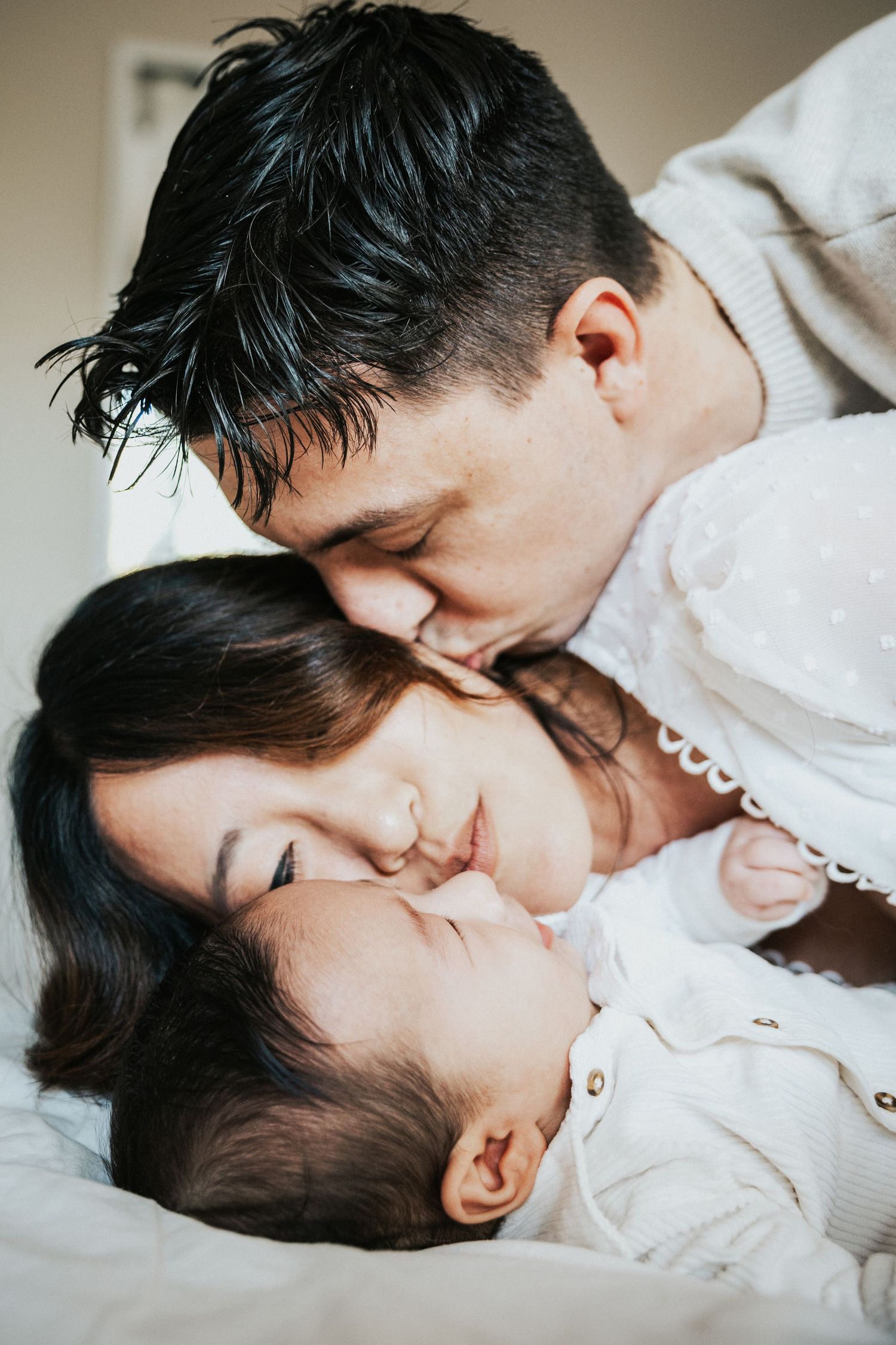 Loving parents gently kiss their sleeping newborn baby while lying together on a white bed.