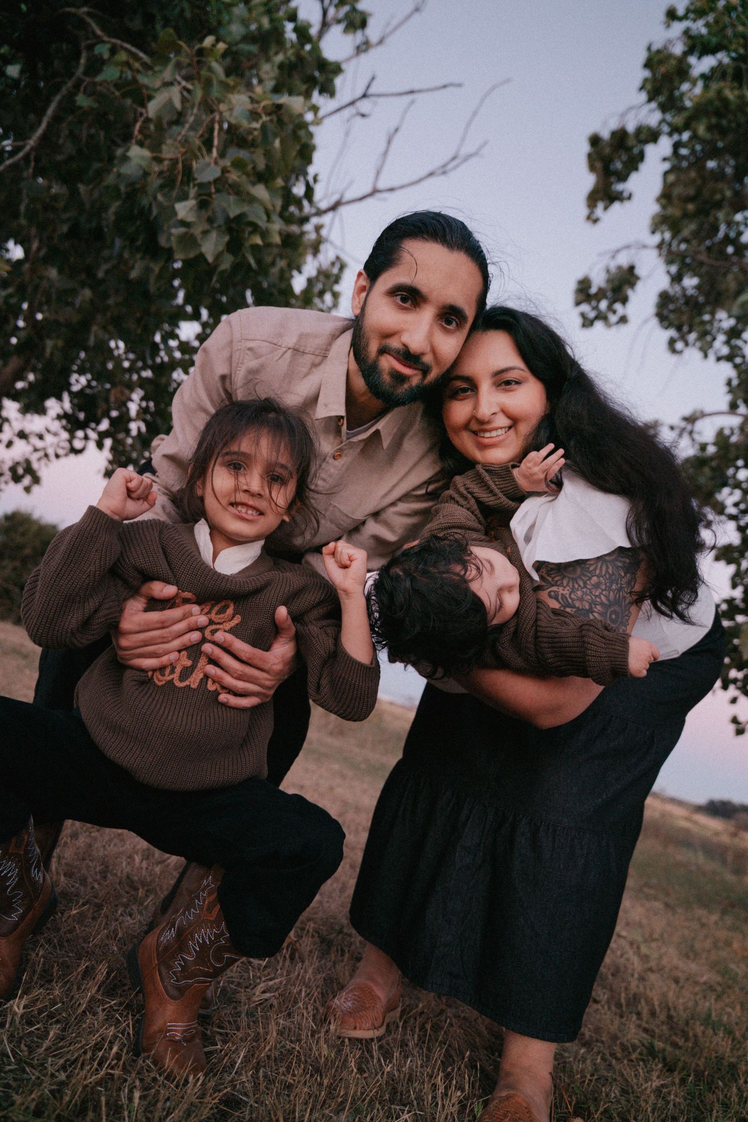 A happy family of four poses outdoors under a tree at dusk, parents hugging their two young children.