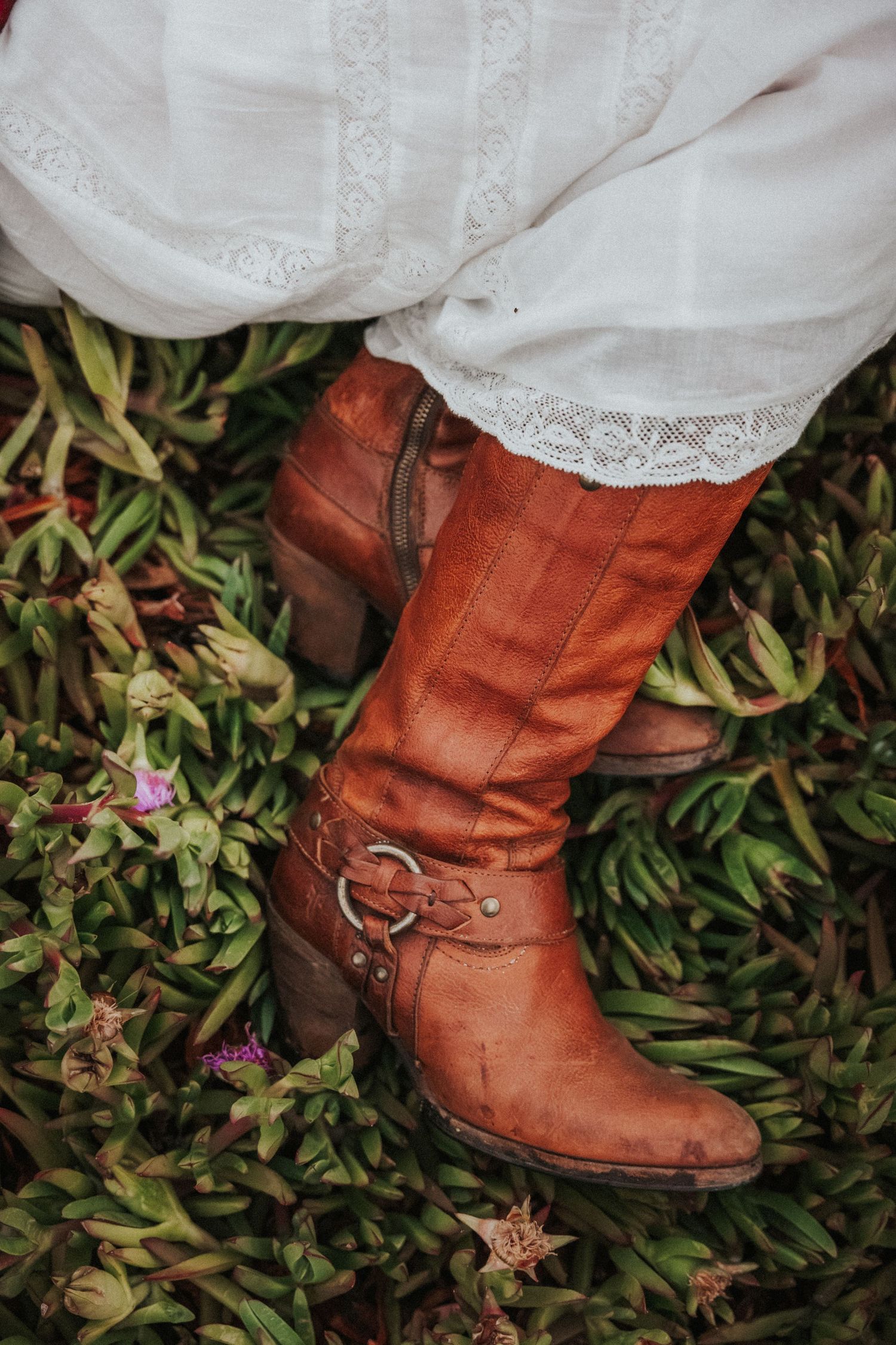 Woman wearing brown leather boots with buckle detail and white lace dress standing on grass with flowers.