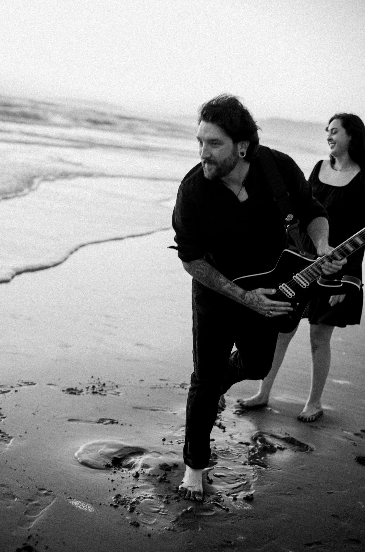 Black and white photo of a tattooed man playing electric guitar on a beach with woman smiling behind him.
