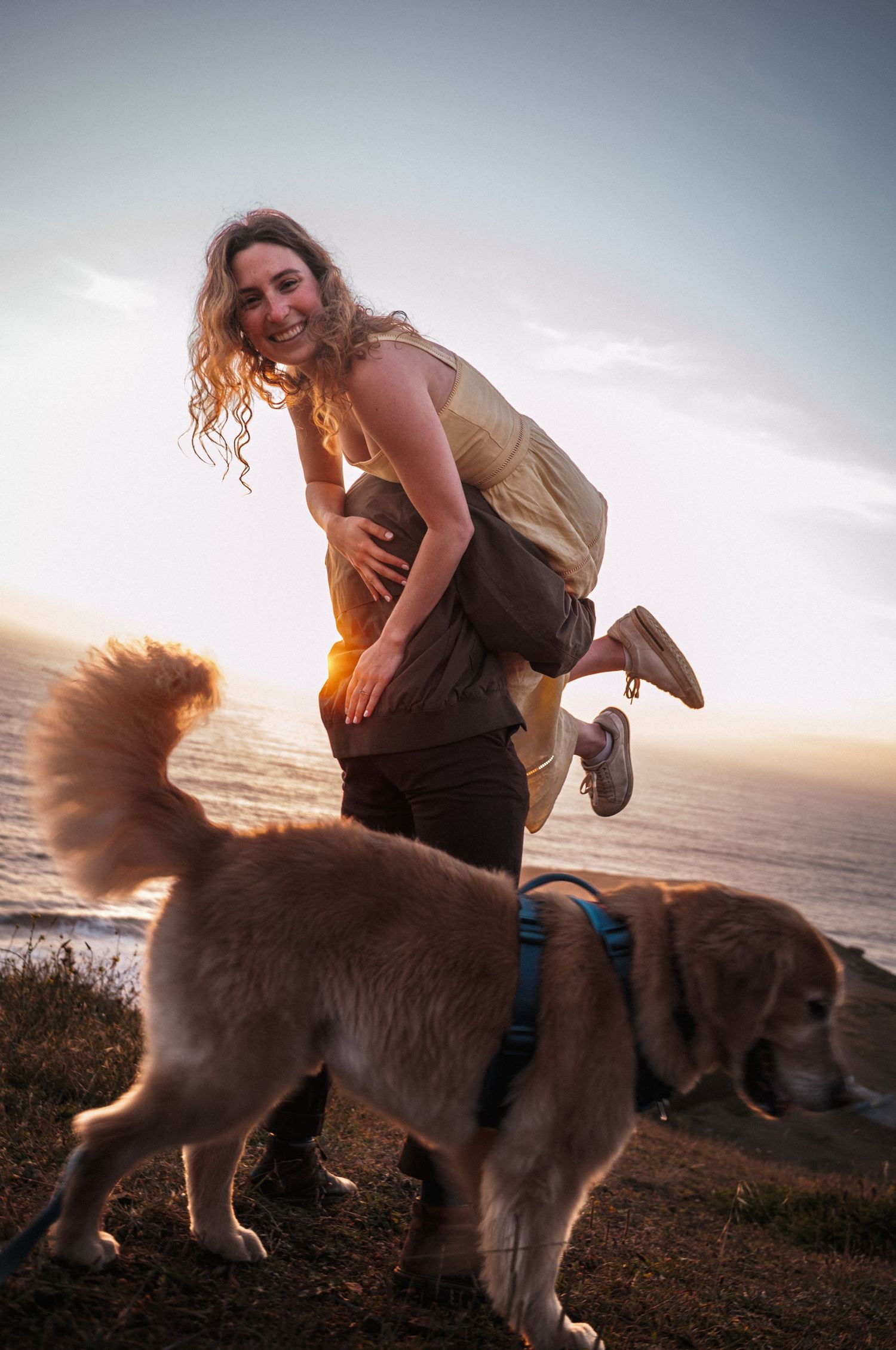 A joyful woman is lifted by her partner near a golden retriever dog at sunset by the ocean.
