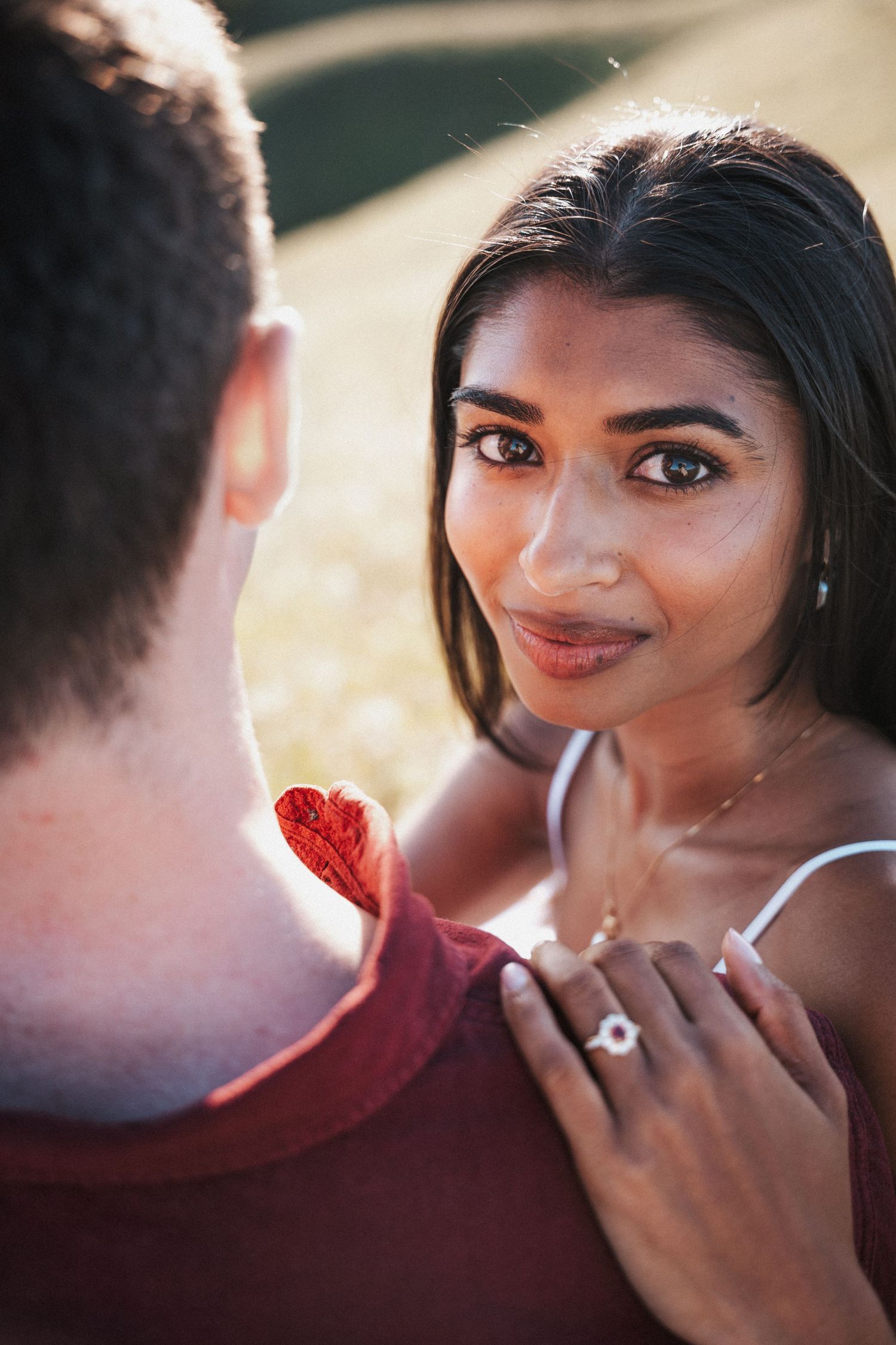 A woman gazes lovingly at a man while holding a red heart, wearing an engagement ring and delicate necklace outdoors.