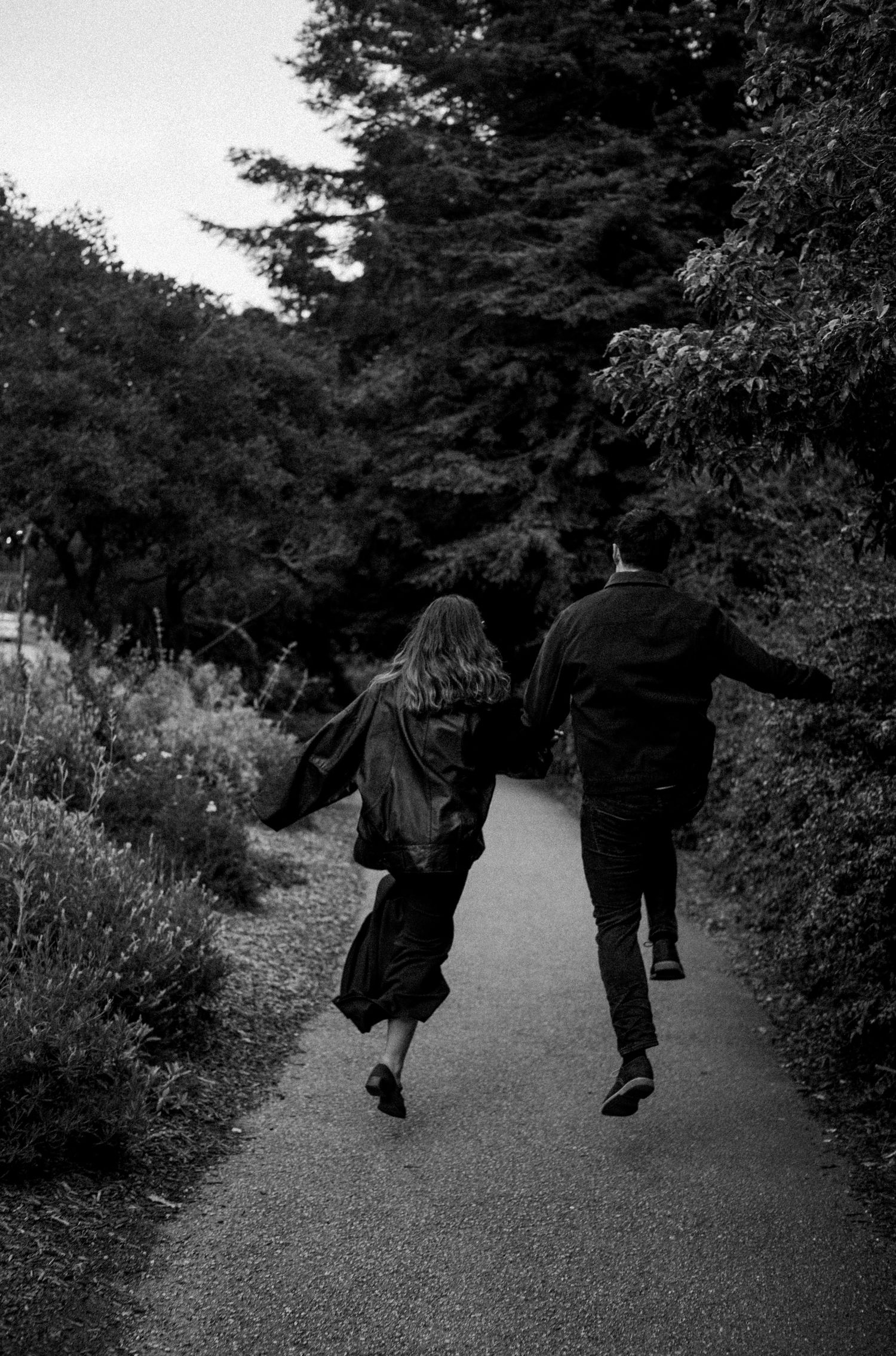 A couple runs hand in hand down a lush garden path in a black and white romantic outdoor photo.
