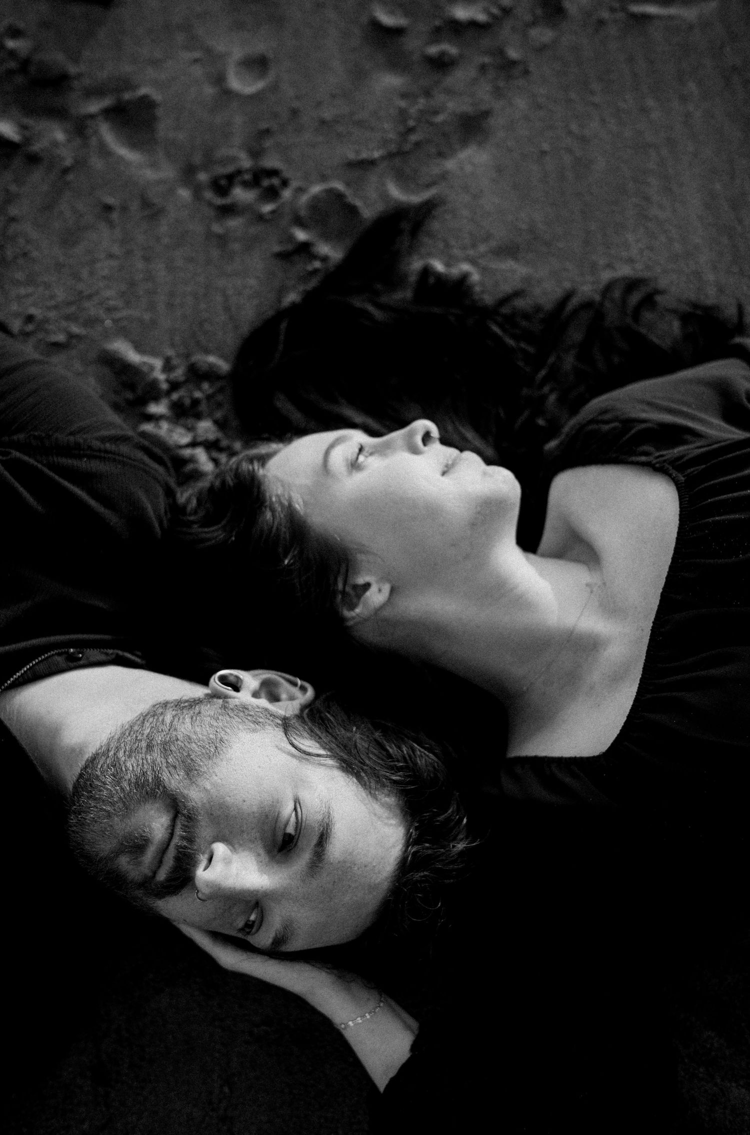Black and white photo of two people lying on dark sand, one looking up peacefully, the other resting eyes closed.Photography by The Y.K. Stu