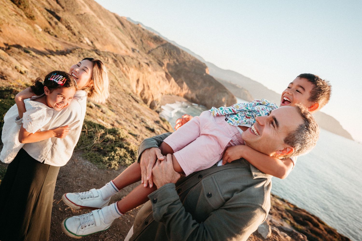Family enjoying a sunny coastal cliffside, with a man carrying a child on his back while a woman smiles nearby.